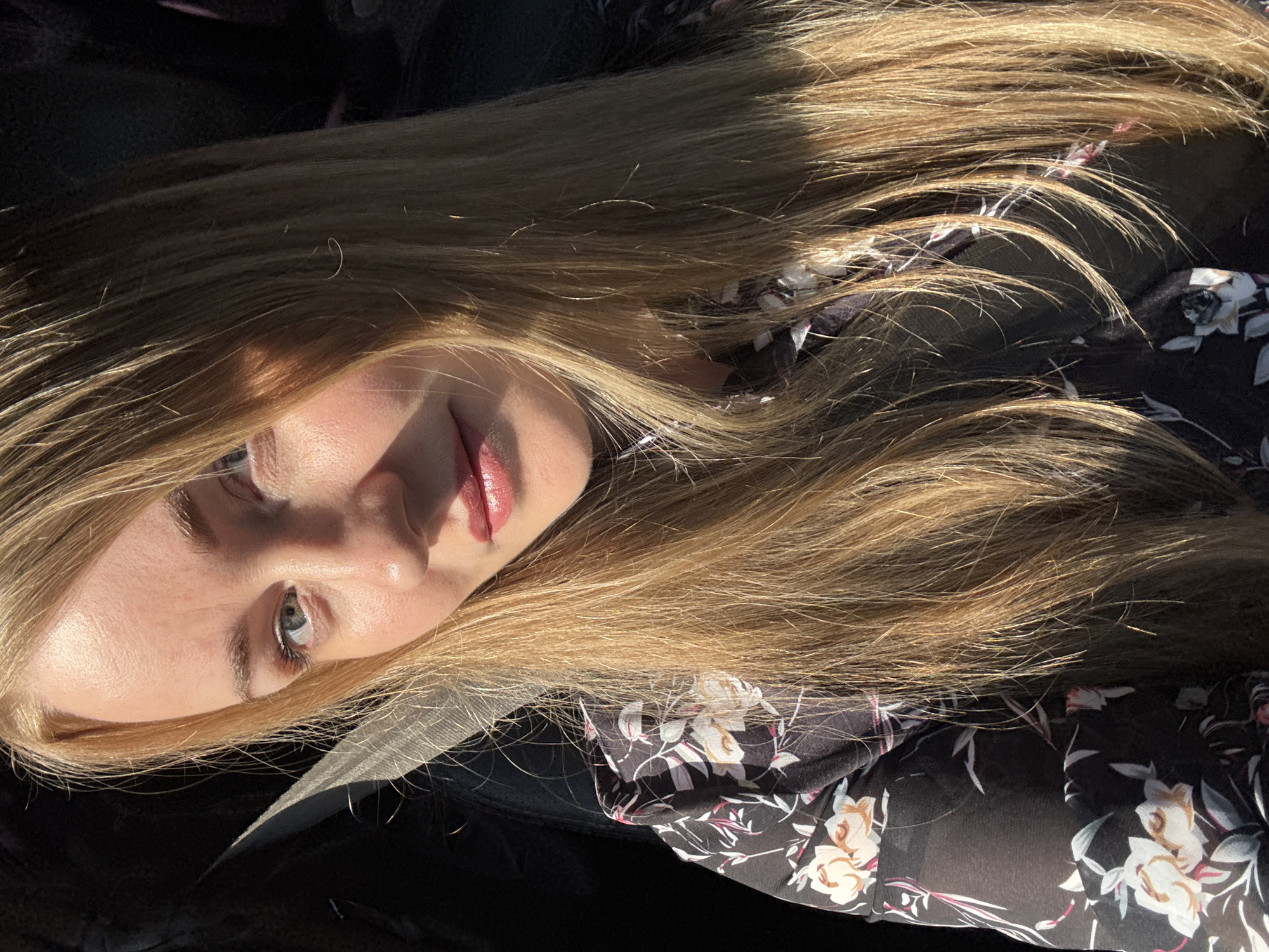 A young woman with long blonde hair and light eyes is reclining in what appears to be a car seat or other dark upholstered surface. She is wearing a black floral print top. The lighting suggests an outdoor setting, possibly Chaikovsky, Russia given the request. There are no explicit visual indicators within the image to definitively confirm the location or activity beyond resting.