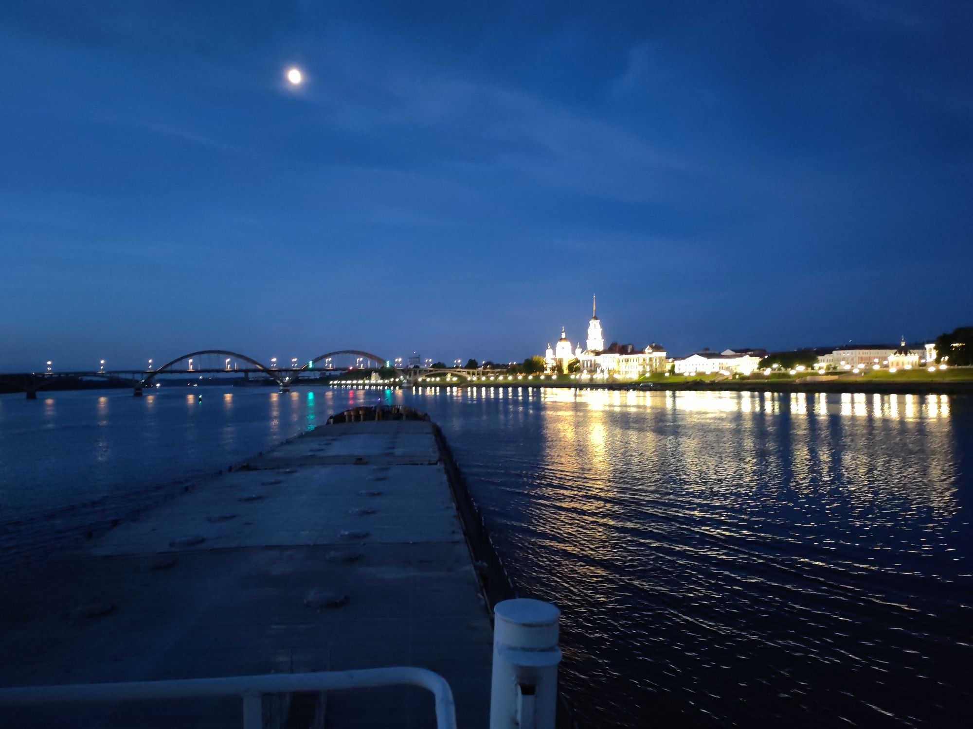 The image captures a nocturnal scene of a wide river with a city skyline in the background. The sky is a deep, dark blue, with a faint crescent moon visible in the upper left quadrant. A multi-arched bridge spans the river in the mid-ground, illuminated by rows of lights. Across the river, a city skyline is visible, characterized by buildings with illuminated windows and architectural details, including spires and domes. The lights from the city reflect shimmering patterns on the dark water's surface, creating streaks of yellow and white light. In the foreground, a concrete structure, possibly a dock or the deck of a vessel, occupies the lower portion of the frame, with a white railing and a white bollard visible. The water in the foreground exhibits gentle ripples, suggesting slow movement. The overall atmosphere is serene and twilight-like, with the city lights providing a warm contrast to the cool blue of the sky and water. No people are visible in the scene.