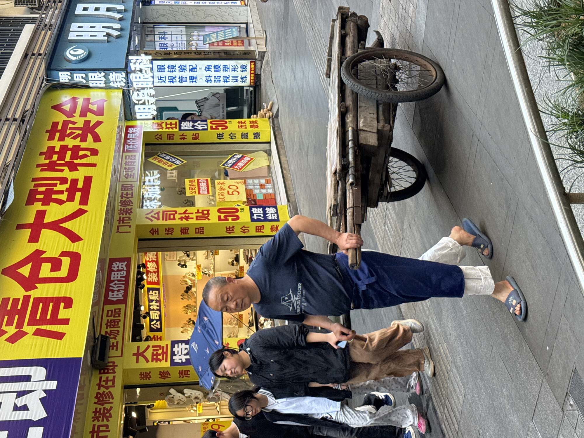 This image depicts a bustling street scene in Jiangjin, China, during what appears to be daytime under indirect or overcast lighting. The central figure is a man, dressed in a navy blue t-shirt with "KEP OUTDOOR" visible and dark blue pants, pulling an old, rustic two-wheeled wooden handcart. He is wearing blue slip-on sandals.
Walking behind him on the grey tiled sidewalk are two women and a child. One woman holds a blue umbrella over her head, while the child, wearing glasses, walks alongside. All individuals are dressed in casual attire.
The background is dominated by a row of vibrant storefronts adorned with numerous signs, predominantly in Chinese characters. A large, prominent yellow banner stretches vertically, emblazoned with "清仓大型特卖会" (Clearance Large Special Sale) in bold red text, indicating a significant sale event. Adjacent to and above this, a blue sign for "明月眼镜" (Mingyue Glasses) and "明月眼科" (Mingyue Ophthalmology) is visible, detailing services such as "近视管理斜弱视训练 眼镜验配角膜塑形镜" (Myopia management, amblyopia and strabismus training, spectacle fitting, orthokeratology lenses).
Other signs advertise various promotions, including "疯狂降价 50%" (Crazy Price Drop 50%), "公司补贴" (Company Subsidy), and "全场商品 低价甩卖" (All items low price clearance). The phrase "市装修在即" or "门市装修在即" (City/Shop renovation soon) is also visible on several signs, likely explaining the clearance sales. The scene captures a typical urban commercial environment with ongoing sales and daily life.