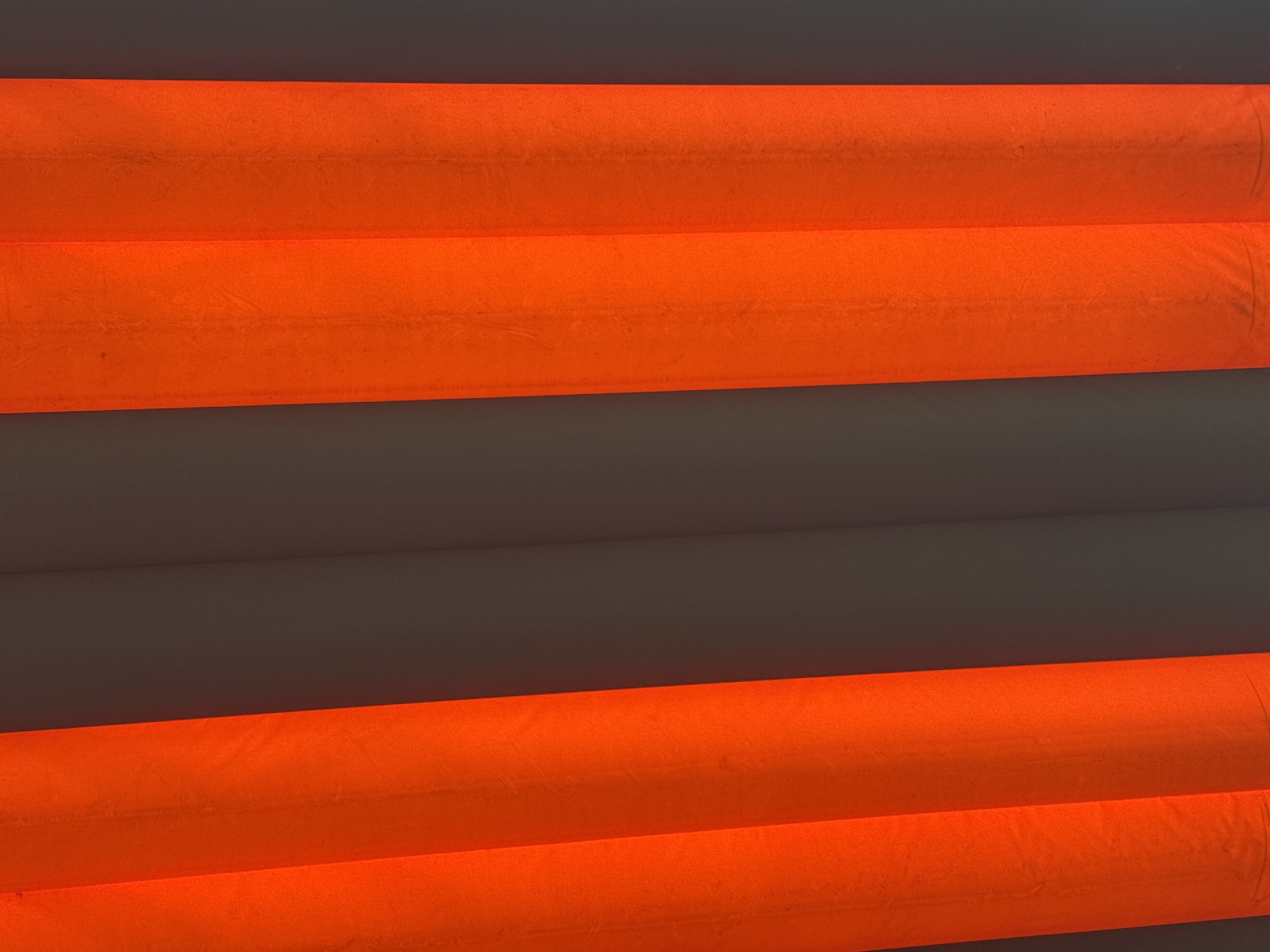 The image displays a close-up, abstract view of what appears to be an outdoor lighting installation. Two vertical elements, glowing with an intense orange-red light, dominate the frame. These illuminated structures have a textured, fabric-like appearance and are separated by a dark, shadowed area. The background, also dark and featureless, suggests a dimly lit or nighttime environment. There are no visible people or discernible urban features that would indicate a specific location in Ia Hru, Vietnam. The perspective is from below, looking upwards, emphasizing the scale and glow of the orange-red elements. The overall impression is one of bold color and abstract form.