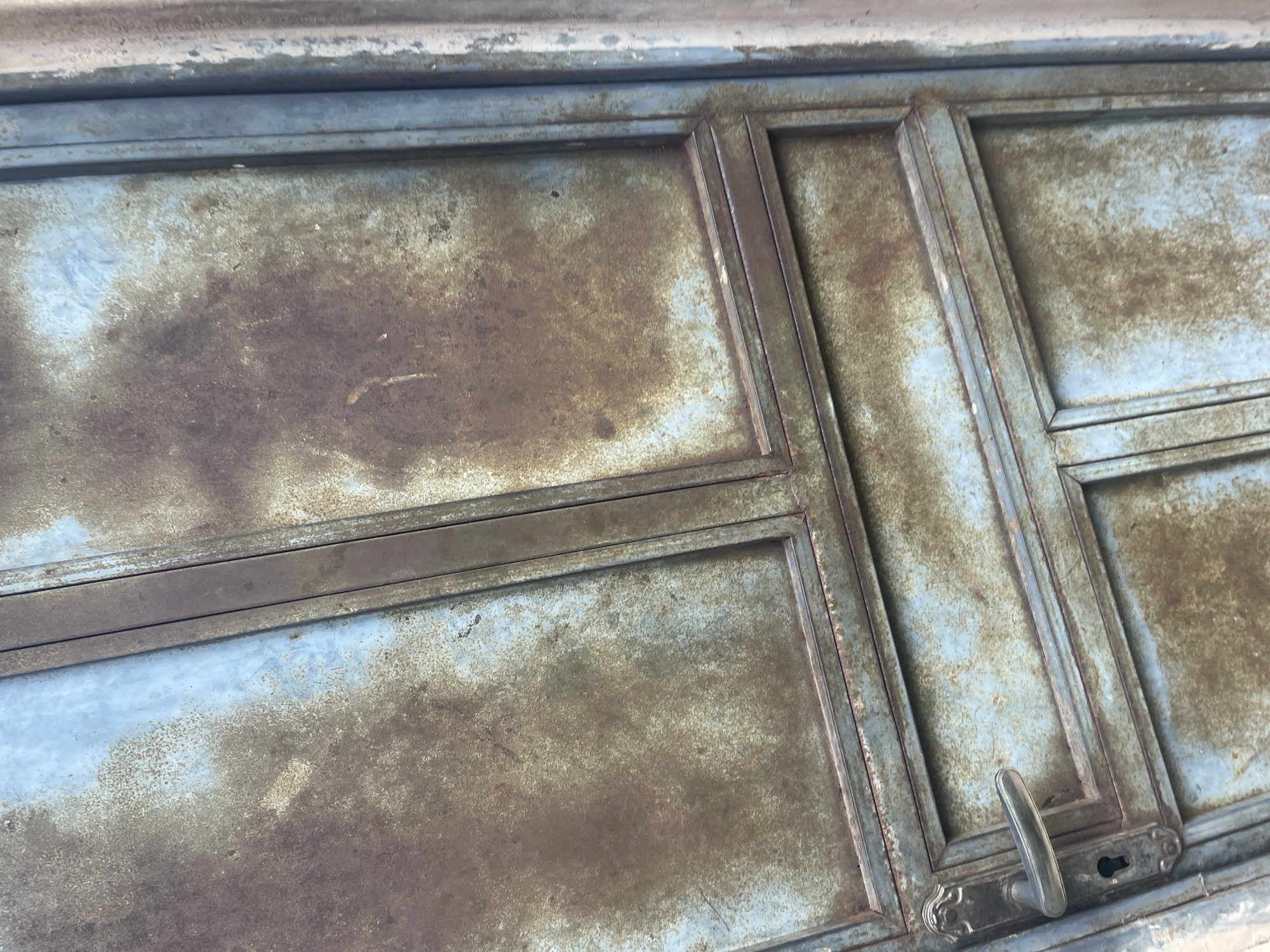 A close-up shot captures the weathered surface of a blue metal door in Lokoja, Nigeria. The door is divided into panels by metal framing. Extensive rust and a patina of dirt cover the blue paint, creating a textured, uneven appearance. In the lower left, a silver-colored door handle and a keyhole are visible. The image has a vertical orientation, with the door occupying most of the frame and a sliver of a beige, textured wall visible on the far right. The lighting suggests an overcast or shaded condition.