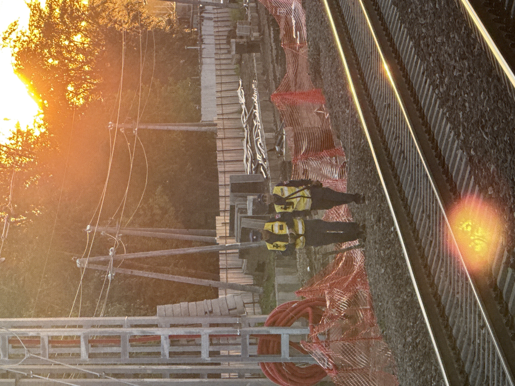 Two individuals in yellow high-visibility vests and dark pants stand near railway tracks. One person is partially obscured by the other, and both appear to be facing away from the camera. The scene is set outdoors during what appears to be either sunrise or sunset, given the warm, golden light casting long shadows and the sun's position low in the sky, partially obscured by trees. The background shows more trees and what looks like utility poles with wires. The foreground features the railway tracks, ballast, and some orange safety netting. A coil of red cable is visible to the left of the netting. The overall impression is of a construction or maintenance site, possibly related to the railway. There is a lens flare in the upper left corner, and a bokeh effect in the lower right. The image's orientation is rotated 90 degrees counter-clockwise. The location is identified as Moscow, Russia.