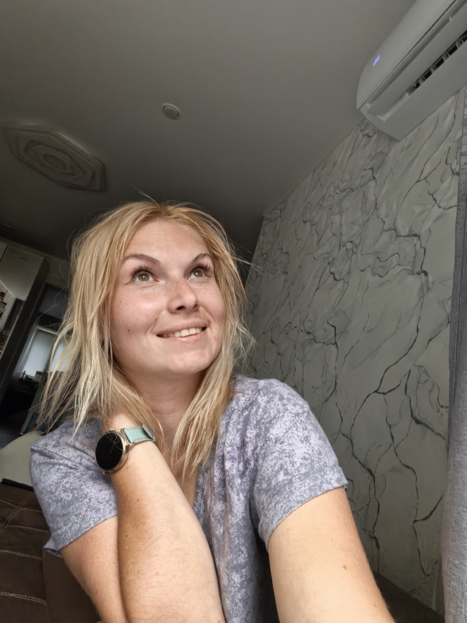 A candid, medium-shot photograph shows a young woman with blonde hair, freckles, and green eyes. She is looking upwards and to her right, with a slight smile on her face. She is wearing a grey t-shirt and a smartwatch with a light blue band. The background consists of a marbled wallpaper pattern on the right and a ceiling with a ventilation grate on the left. An air conditioner unit is visible on the wall in the upper right corner. The lighting suggests it is daytime.