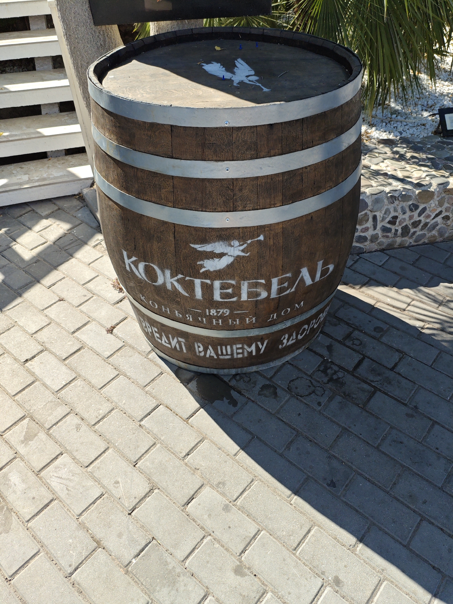 A weathered wooden barrel with metal bands is positioned outdoors on a paved surface. The barrel has white lettering that appears to be in Russian, with the word "КОКТЕЙЛЬ" prominently displayed. There is also a depiction of a winged figure on the barrel. The barrel is set against a backdrop that includes white stairs, some greenery, and a wall constructed of stones. The lighting suggests it is daytime, with shadows cast on the pavement. The overall scene is likely from a public outdoor area, possibly a courtyard or garden. There is no indication of the city of Alushta, Ukraine within the image itself.