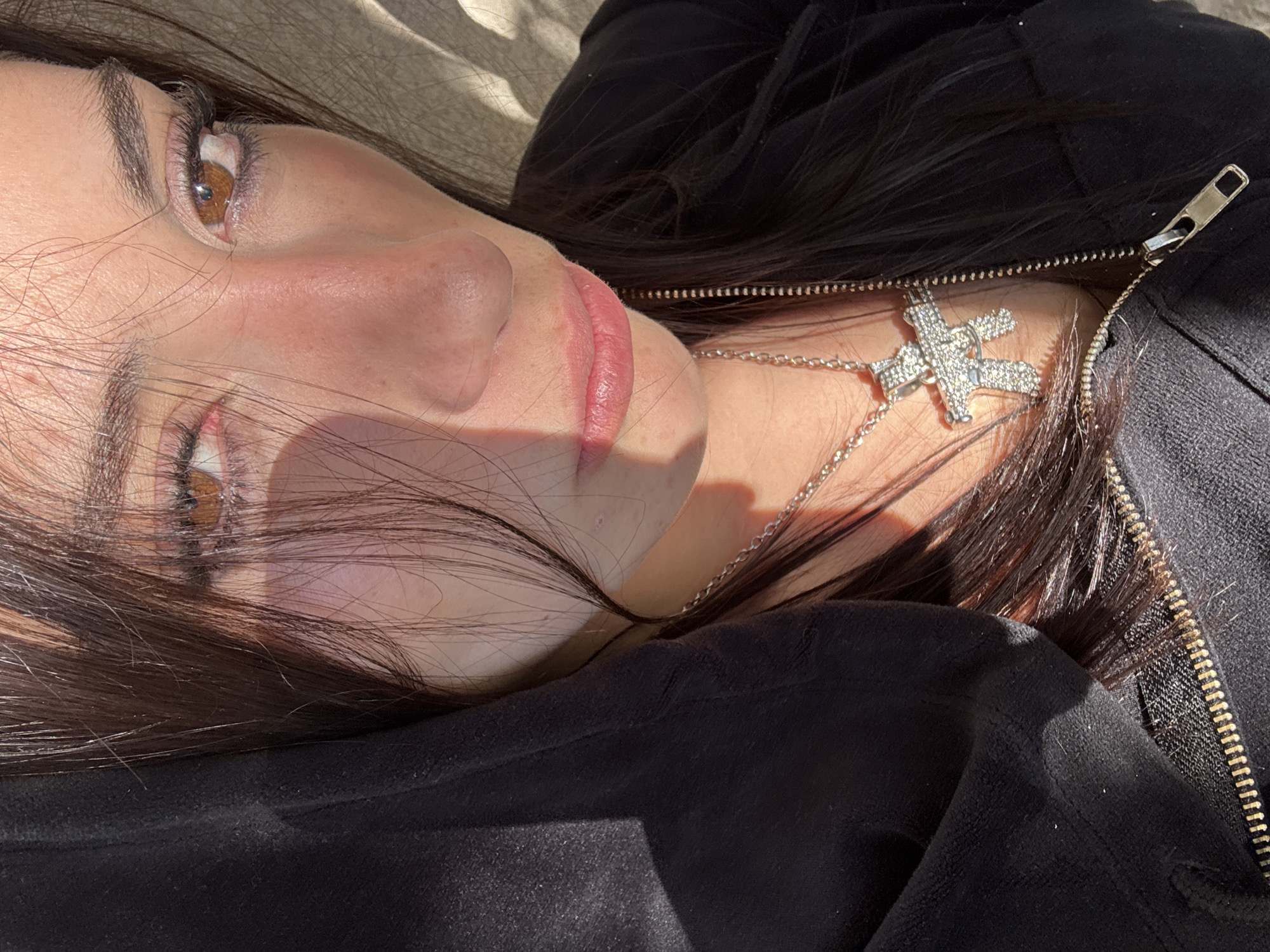 A young woman with light skin and brown eyes is lying down, possibly outdoors, wearing a black zip-up hoodie and a silver necklace with a "K" pendant. Part of her face is shadowed by the sun. The location is not evident from this image.