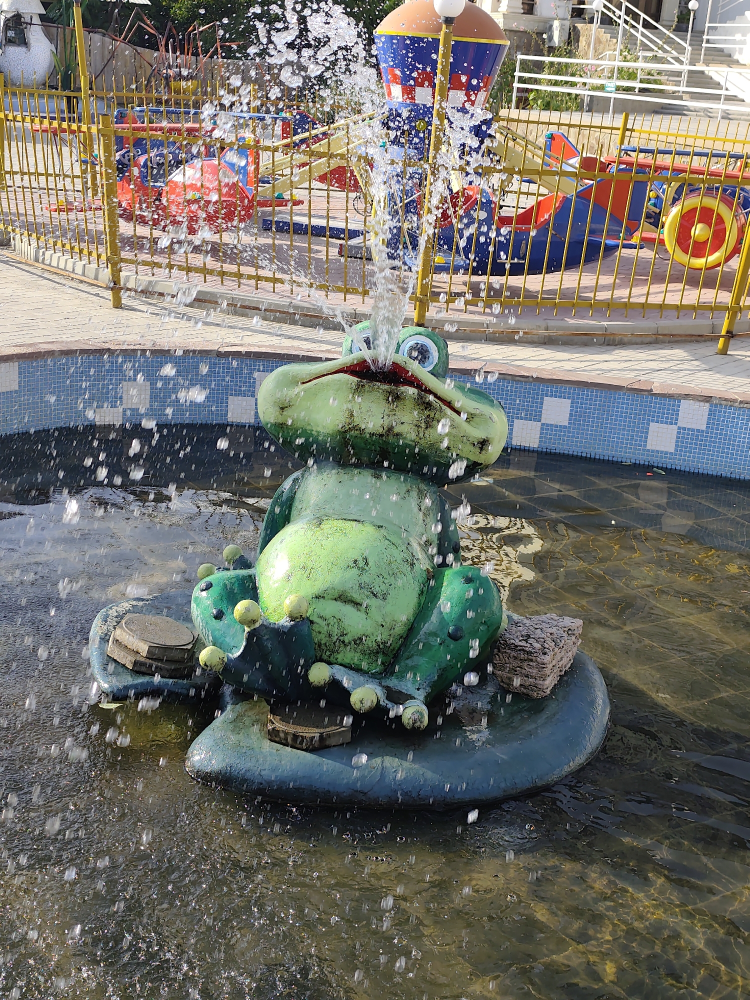 A green frog fountain sprays water in a shallow pool. The fountain is in the foreground, with a yellow metal fence and a playground in the background. The playground features colorful rides and structures, including a rocket-shaped object. The ground around the pool is paved with light-colored tiles. The scene appears to be outdoors in daylight.