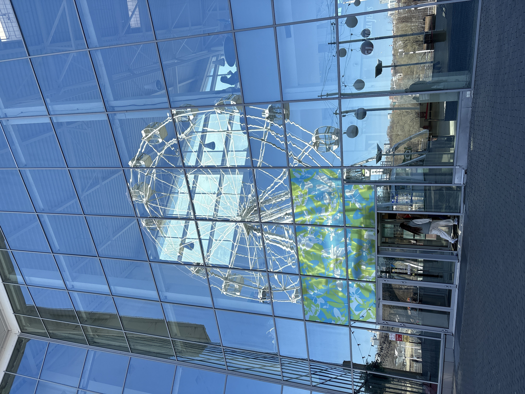 The image shows a building with a glass facade reflecting various objects. Reflected in the glass are the sky, part of a metal Ferris wheel structure, street lamps, and part of a landscape with trees.
Near the entrance, the reflection shows a woman in light-colored clothing walking through the doorway. The facade also features decorative panels with blue and green floral patterns near the entrance.
The location is Ulyanovsk, Russia.