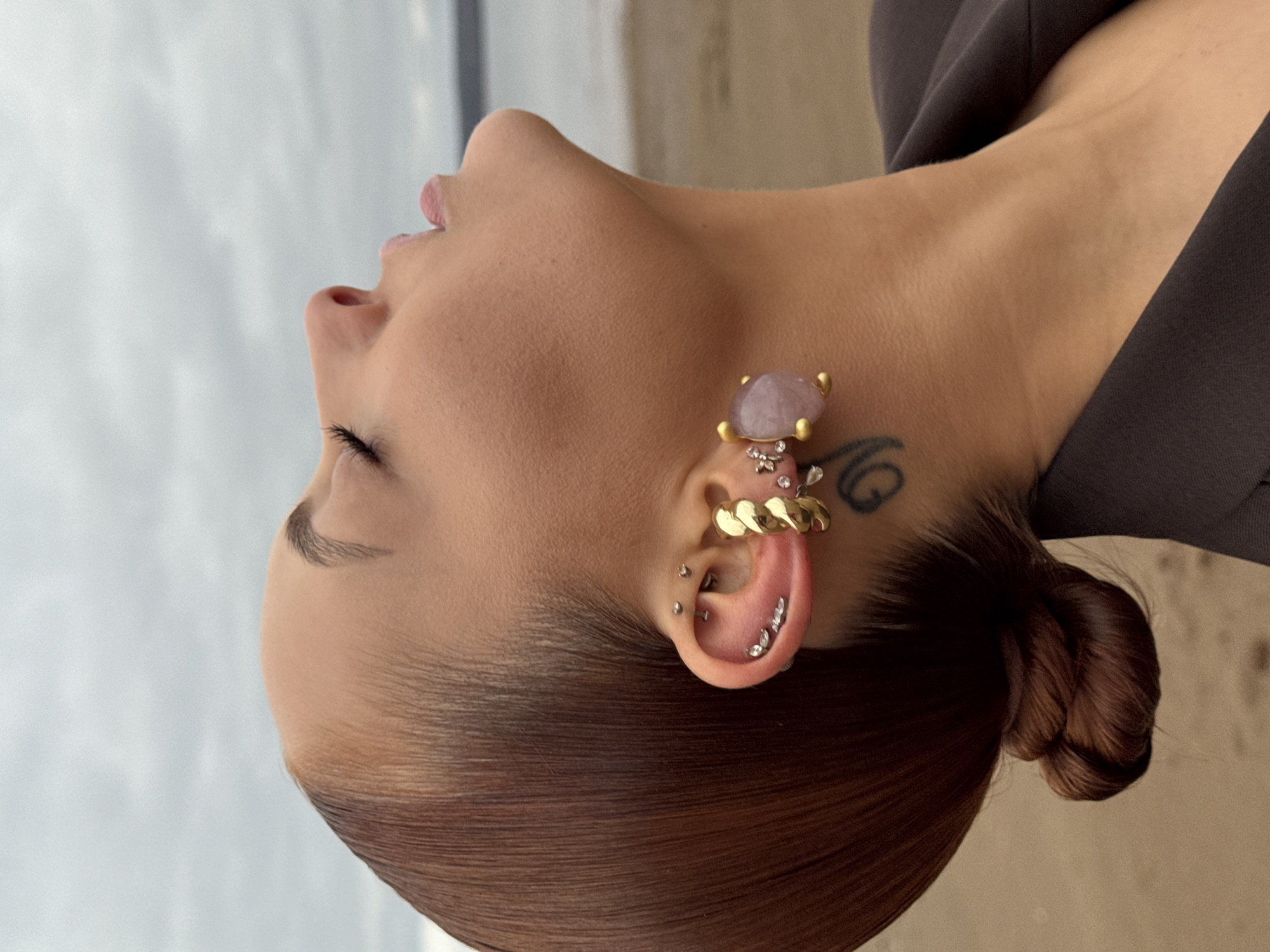 The image shows a woman from the shoulders up, tilting her head back towards the sky. She has fair skin and brown hair pulled back into a bun. Her eyes are closed, and her lips are slightly parted. She is wearing a dark brown top. Her ear is adorned with multiple piercings and jewelry, including a gold ear cuff, a row of small studs, and a larger decorative earring with a pink stone. A tattoo is visible near her ear. The background appears to be a beach with sand and a cloudy sky. There is no information indicating the photo was taken in Chaikovsky, Russia.
