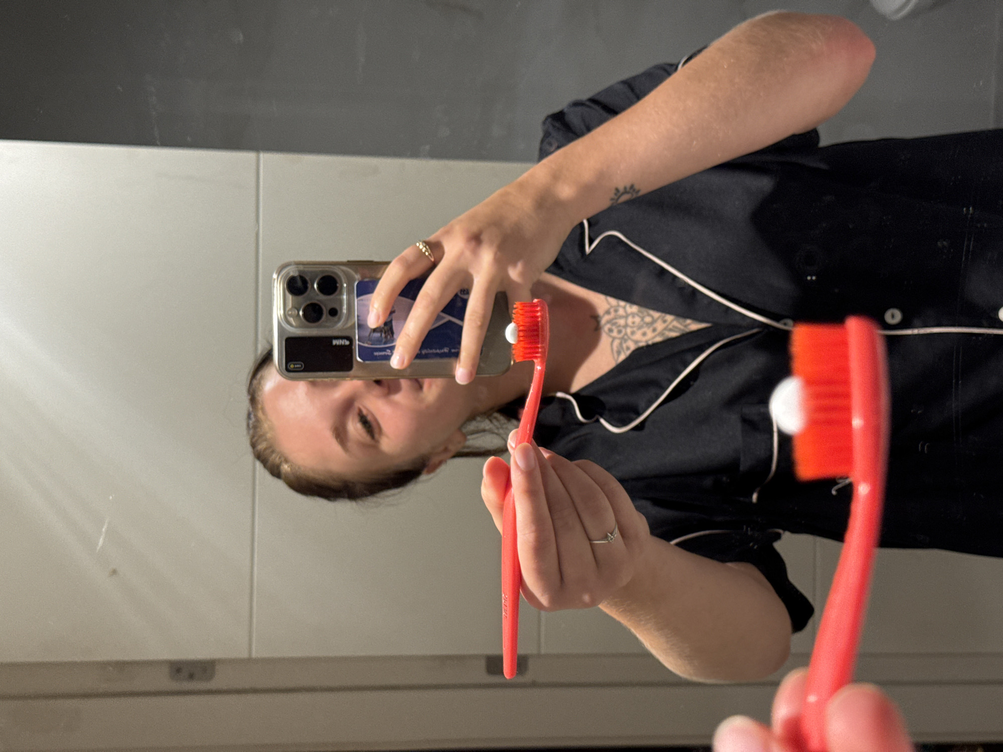 A young woman is taking a selfie in a bathroom mirror, holding a red toothbrush with toothpaste on it. She is wearing black pajamas with white trim and has tattoos on her chest and arm. The bathroom appears to be modern with gray tiles and a large mirror. The woman is smiling and looking at her phone. She is in Aksu, Turkey.