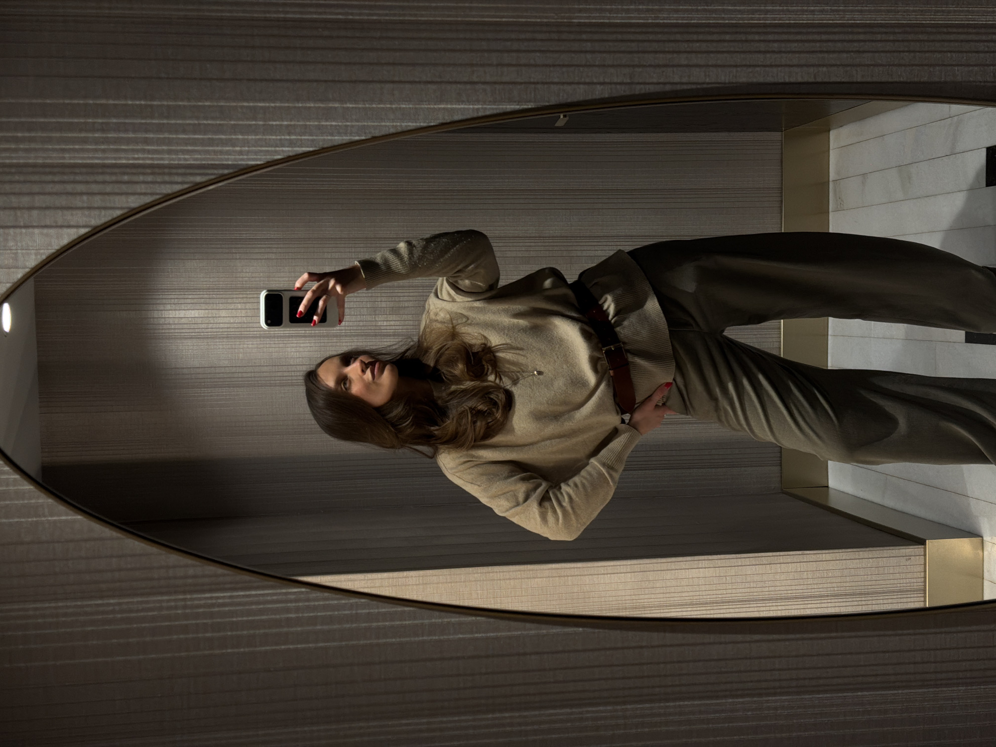 A woman is taking a selfie in a mirror. The mirror is oval-shaped with a bronze-colored frame. The woman has long brown hair, and she is wearing a beige sweater, brown belt, and olive-green pants. She is holding a smartphone in her right hand with red nail polish on her nails. The background reflected in the mirror shows a wall with horizontal stripes and a floor with white marble tiles. It is possibly a luxury hotel bathroom, as the picture may have been taken at the Dukley Hotel & Resort in Budva, Montenegro.