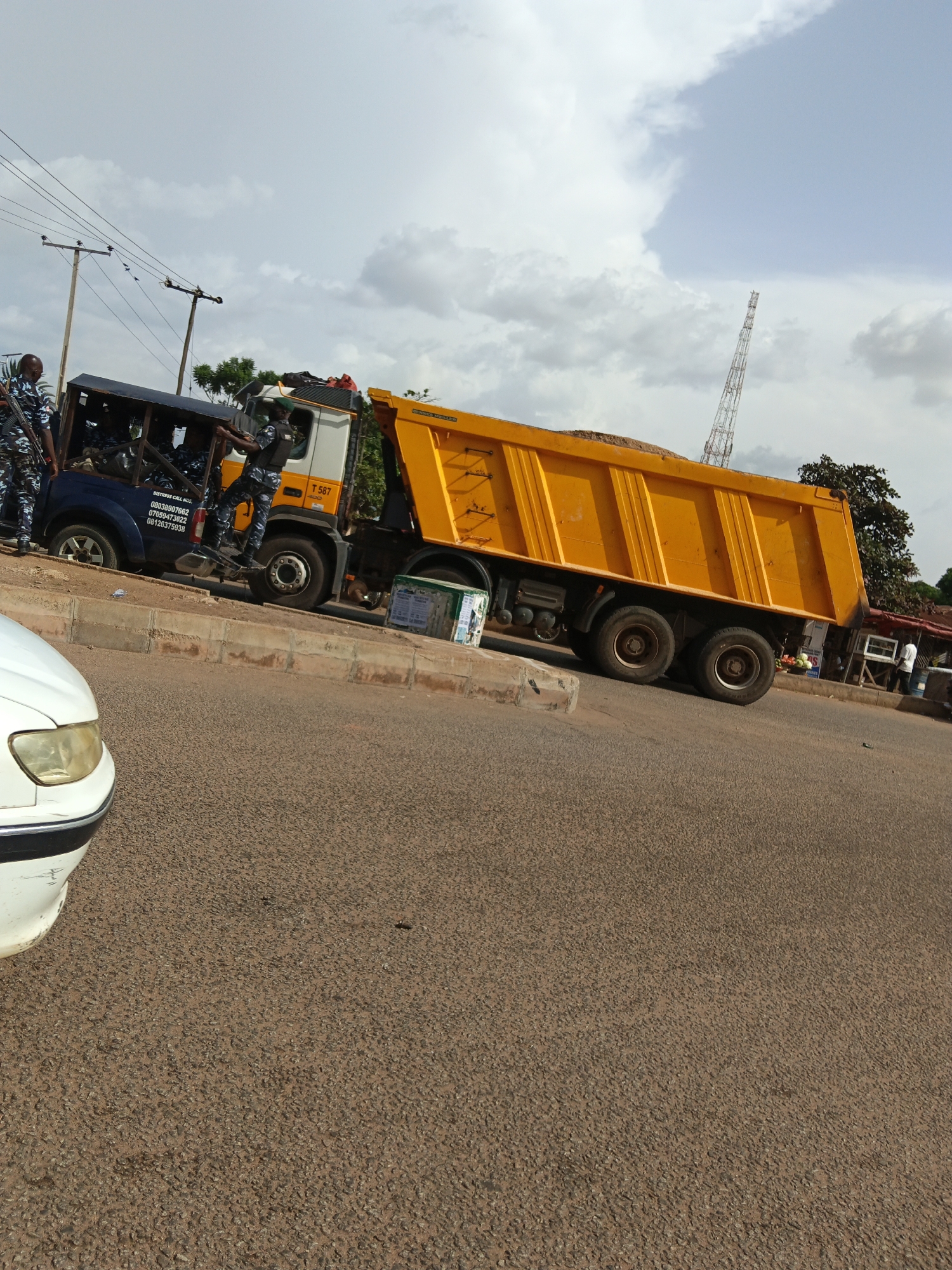 In an outdoor scene, a bright yellow dump truck is parked on a roadside, its bed filled with sand. The truck has a white cab with "T 587 400" printed on its side. Several individuals, appearing to be law enforcement officers in camouflage uniforms, are positioned around the truck. Two officers are standing on the side of the truck's cab, and several more are visible inside the blue truck bed behind the cab. A white car is partially visible in the foreground, on the left side of the image, facing right. The background features a cloudy sky, utility poles with wires, and some trees and buildings further in the distance. The ground is paved with asphalt.