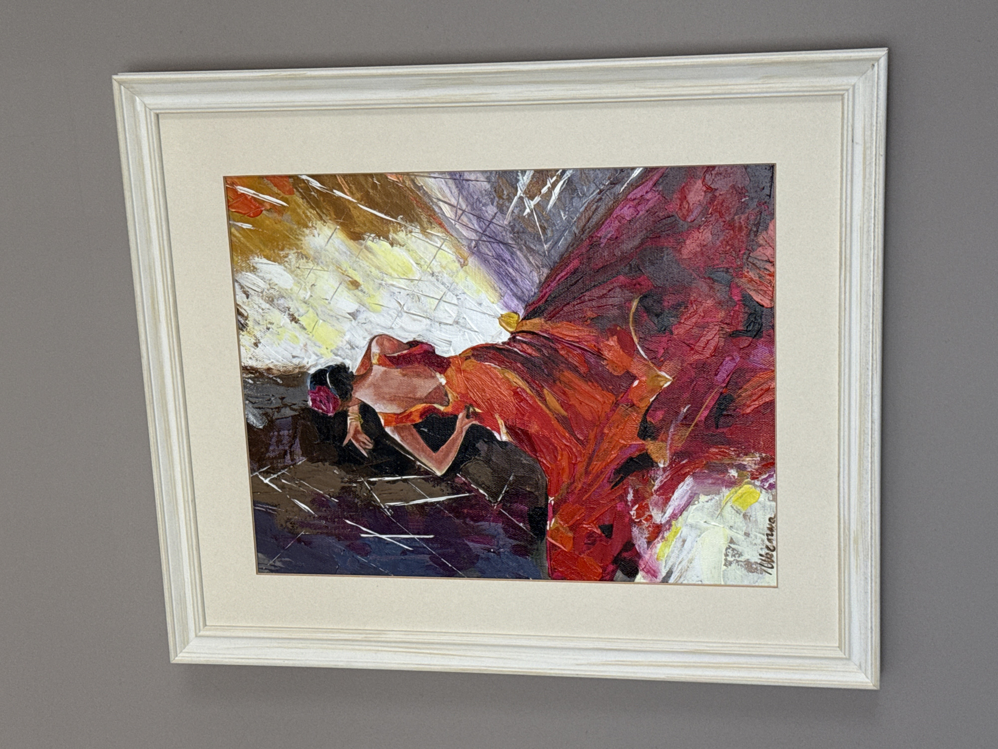 A framed painting of a flamenco dancer is displayed on a grey wall. The painting depicts a woman in a flowing red dress with her back to the viewer, her left hand on her hip and her right arm extended. Her dress is richly textured and voluminous, with a gradient of reds and purples. A pink flower is tucked into her dark hair. The background is a dynamic abstract composition of dark purples, browns, and bright yellows, with visible brushstrokes. The painting is signed in the bottom left corner with a name that appears to be "W. Wierzawa". The frame is a light-colored wood with a slightly distressed finish. The painting is hung on a plain grey wall, suggesting it is in a private residence or gallery. There is no specific indication of the location within Burgas, Bulgaria.