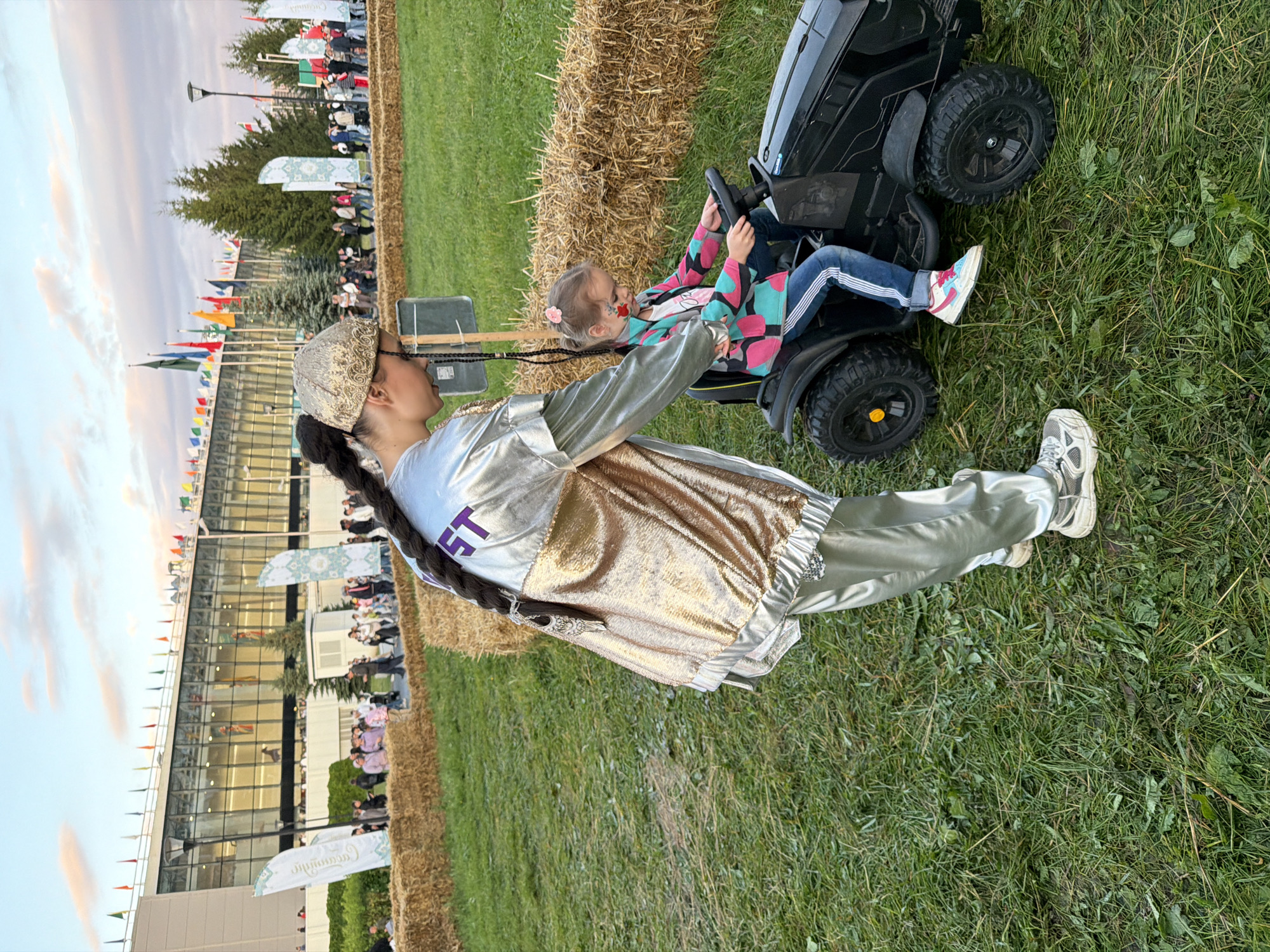 Two females are present outdoors on a grassy area. In the foreground, a young girl is seated in a black toy tractor. She has on a colorful jacket, blue jeans with white stripes, and sneakers. Her hair is pulled back, and she has some face paint on. A taller female stands next to the tractor, with her arm on the girl's shoulder. She is wearing a shiny silver tracksuit and a gold patterned head covering. Her hair is braided down her back. Behind them, a large building with many windows is visible, with flags displayed on its roofline. Several other people are walking in the background, near the building. The ground is covered in grass and hay bales are arranged as a barrier. The sky is cloudy and appears to be late afternoon or early evening.