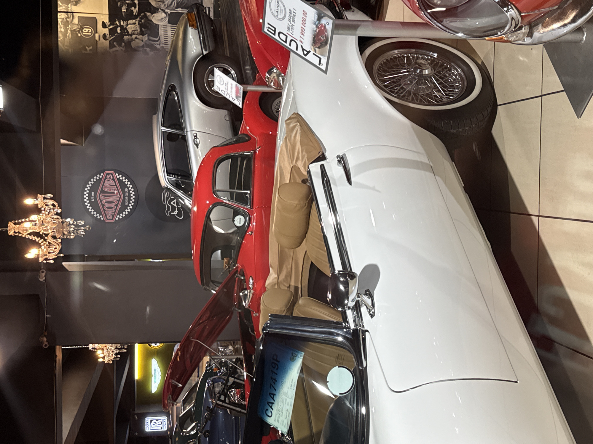 The image shows a car dealership or showroom with several classic cars on display, likely located in Cape Town, South Africa (based on the "CAA" prefix on the license plate). In the foreground is a white 1962 Jaguar E-Type Series 1, with a price tag of R1,999,000.00. Next to it is a red classic car and a silver car in the background. A sign for "BROOKLANDS MOTOR CO LTD" is visible on the wall. A Rolls Royce sign is also visible. Chandeliers are hanging from the ceiling. The image is well-lit and focused.