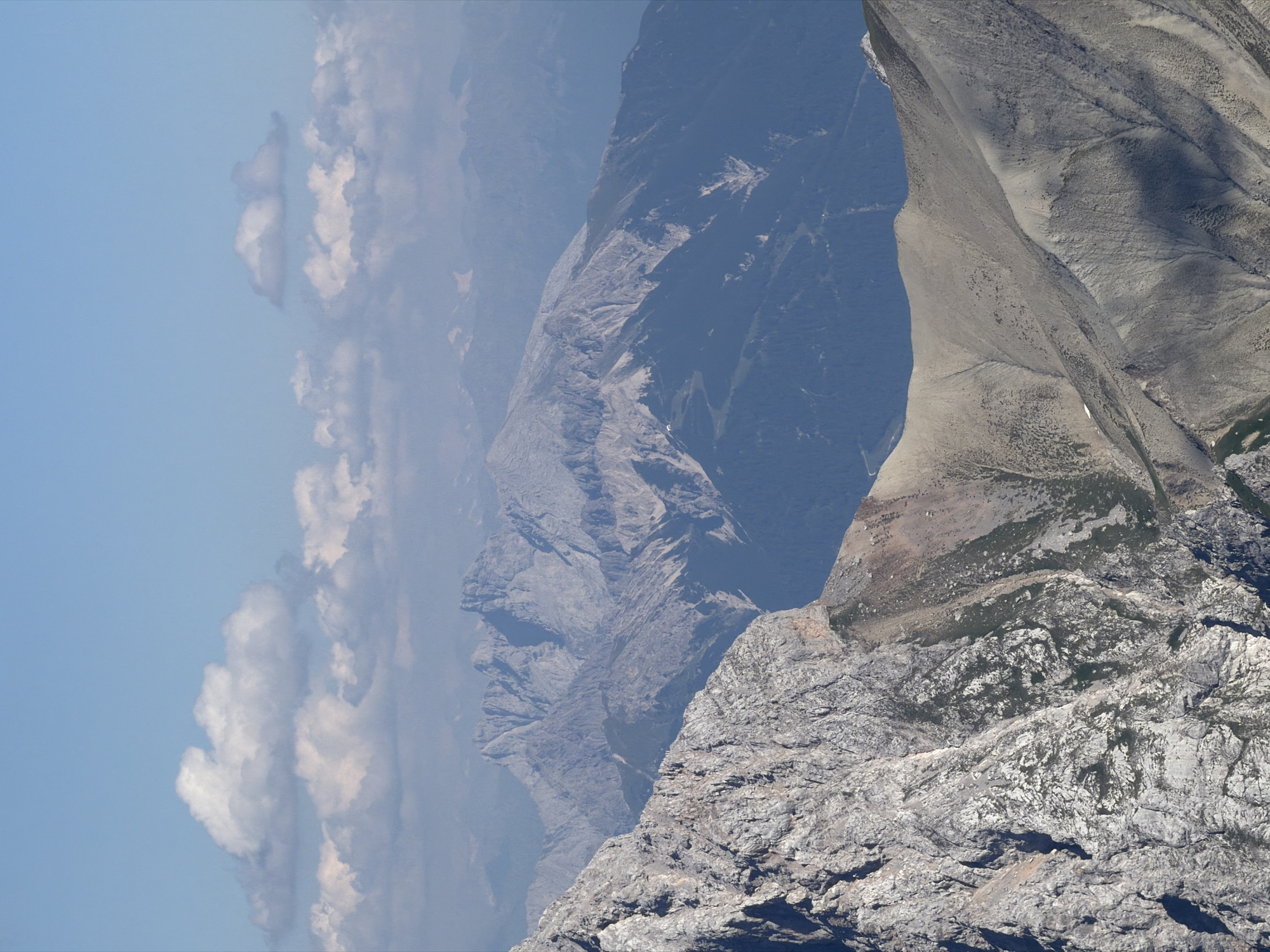 The image shows a panoramic view of a mountainous landscape, likely taken from a high vantage point. The foreground features a jagged, rocky slope with visible cracks and texture. In the midground and background, a series of majestic, rugged mountains stretch across the frame, displaying varying shades of gray and bluish hues due to atmospheric perspective. Patches of dark green vegetation, likely coniferous trees, are visible on the lower slopes of some mountains. The sky above is a clear, pale blue, dotted with scattered white, fluffy clouds. The scene evokes a sense of wilderness and natural grandeur. This landscape is reminiscent of the Bavarian Alps, which are in the vicinity of Garmisch-Partenkirchen, Germany, a well-known alpine resort town.