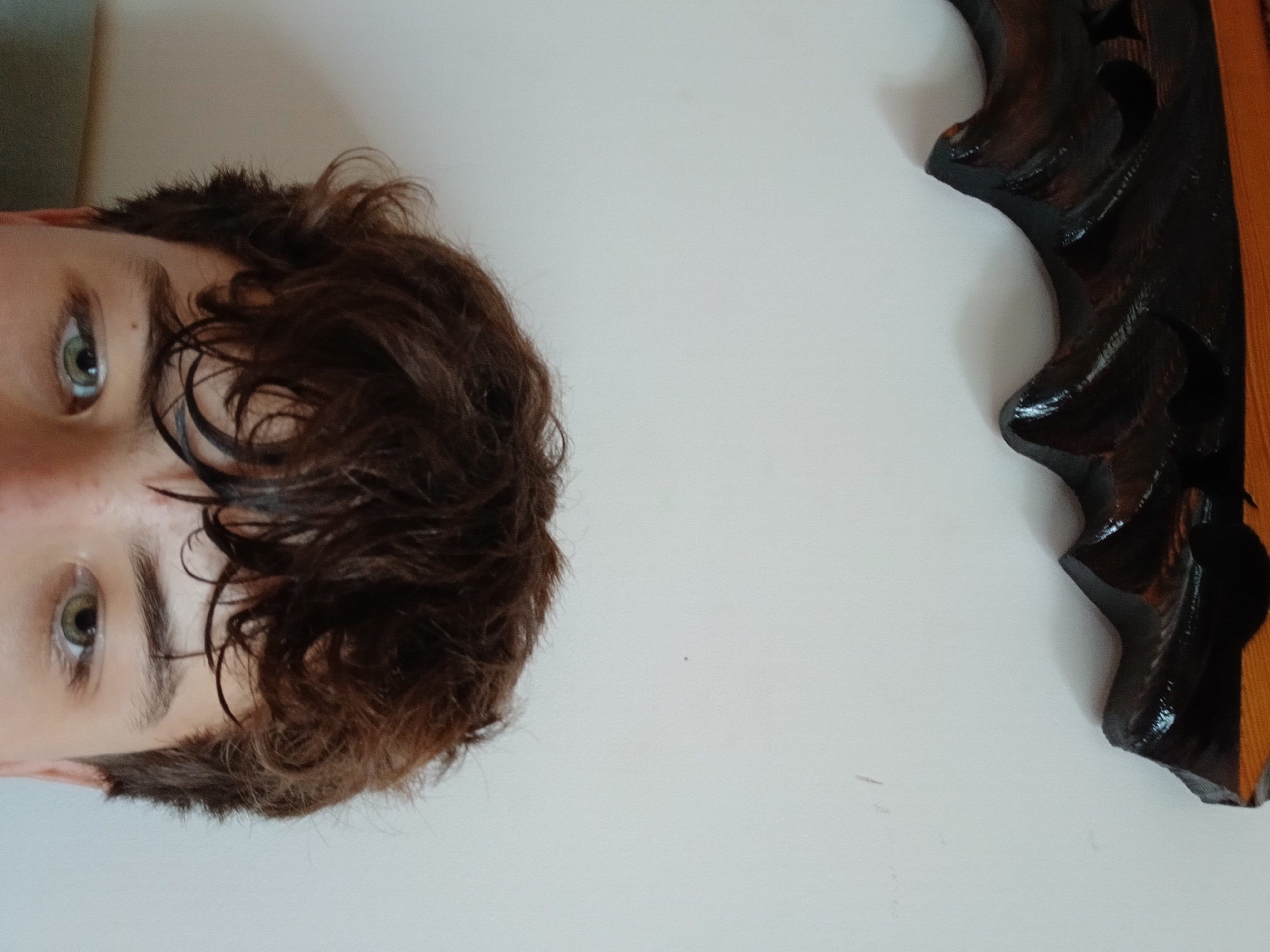 The image shows a close-up view of a person's face, focusing on their eyes and brown, curly hair. The person's left eye, which is green, is clearly visible. The background consists of a plain white wall with a dark, ornate wooden object at the top of the frame. No specific location within the city of Dnipro, Ukraine, can be identified from the provided image.