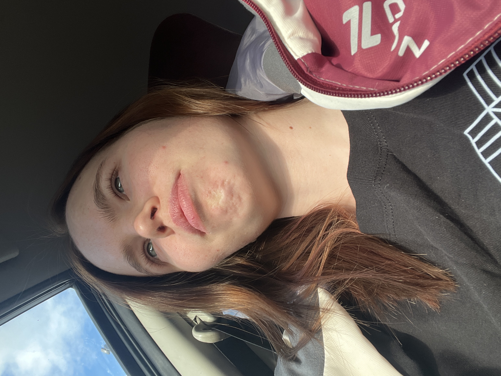 A tilted medium shot captures a young woman inside what appears to be a car, looking directly at the camera. She has long, reddish-brown hair that cascades over her shoulders and against a dark t-shirt. Her eyes are a light color, possibly blue or green, with full eyebrows. Her expression is neutral and calm, perhaps slightly pensive. Visible on her skin are natural textures, including pores and some minor blemishes or marks on her cheek and chin area.
She is wearing a dark, possibly black, t-shirt. Over her right shoulder (viewer's left), part of a red and white zippered item, resembling a jacket or bag, is visible. White text on this red fabric clearly shows "1L" followed by stylized characters that could be interpreted as "%" or "PN".
The setting is an automobile interior, with a bright car window to her left revealing a clear blue sky dotted with white clouds, indicating it is daytime with fair weather. The interior of the vehicle is otherwise dark, with natural light illuminating her face from the side. The overall scene suggests a candid moment within a vehicle in Russkie Kirmeni, Russia.