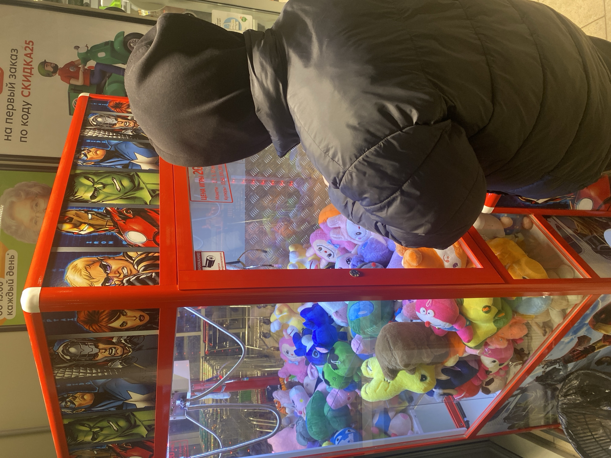 A person wearing a dark hooded jacket is bending over a claw machine in what appears to be a store in Votkinsk, Russia. The claw machine is red and decorated with Marvel superhero characters on the side. The machine is filled with various colorful plush toys. A sign next to the machine advertises a discount for first-time orders, and another sign indicates the game costs 20 rubles and the store practices video surveillance. Other signage in Russian is visible in the background.