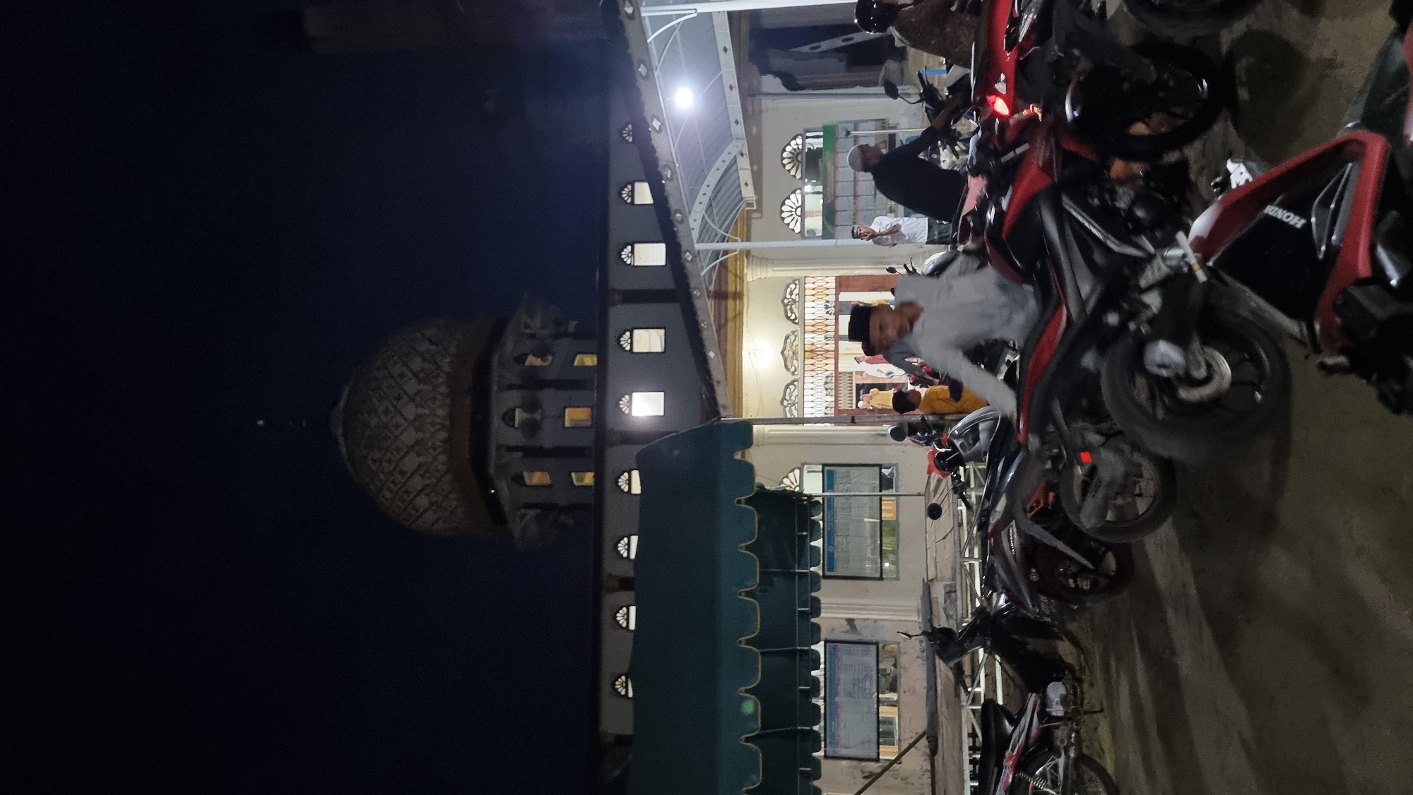 The image depicts the exterior of a mosque at night in Manetunong, Indonesia. The mosque features a prominent, ornate dome illuminated from within. A multi-story facade with arched windows and decorative elements is visible. In the foreground, a collection of motorcycles is parked, some with riders. A few individuals, appearing to be men, are seen in and around the mosque entrance, some wearing traditional Islamic headwear. The scene is lit by artificial lighting, creating shadows and highlighting the architectural details of the mosque and the parked vehicles. The overall atmosphere is that of a community gathering or prayer service taking place at night.