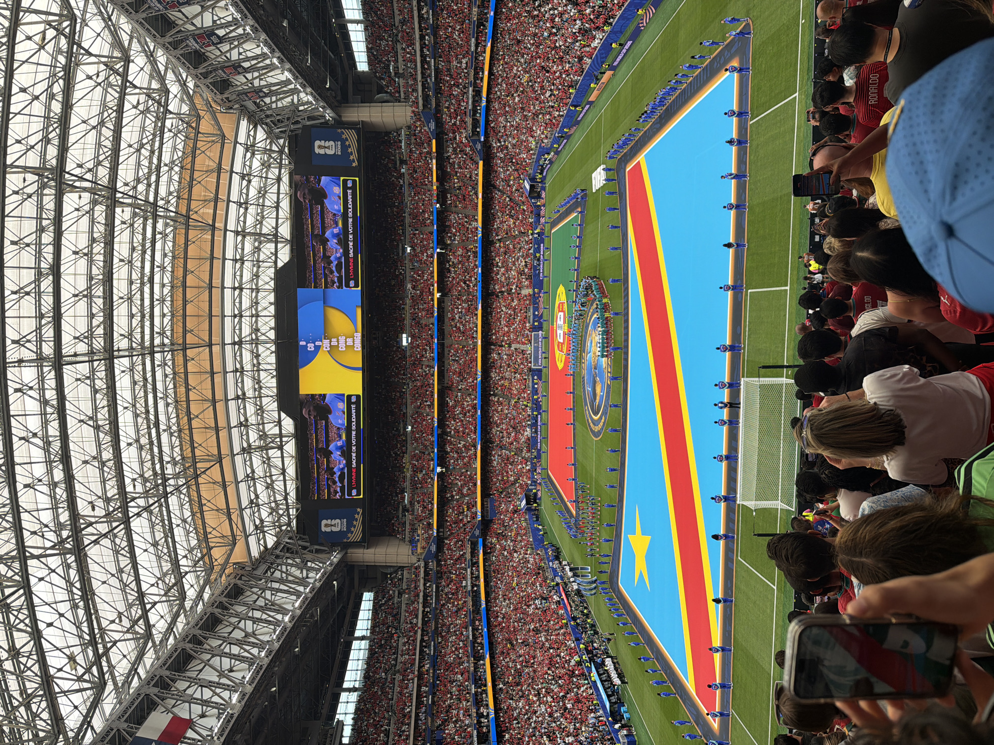 This is a high-angle, wide shot of a soccer stadium during a pre-game ceremony. The main subjects are a large contingent of people, including players lining up on a soccer field and a massive crowd filling the stadium seats. The field itself is dominated by a large replica of the flag of the Democratic Republic of Congo, with its distinct blue, red, and yellow colors, and a yellow star. Players, dressed in matching blue uniforms, stand at attention along the edges of the flag. The stadium is vast and appears to be covered by a large, intricate metal roof structure.
The event taking place is likely the pre-game national anthem or introductions, as suggested by the formal lineup of players and the presence of the flag. The atmosphere seems anticipatory, with the large crowd indicating a significant event. The lighting suggests it is daytime, and the setting is an indoor stadium, likely in Houston, Texas, given the location context.
Notable details include the large video screens visible at one end of the stadium, displaying images and text. One screen shows a close-up of what appears to be a soccer team and text in French: "L'HYMNE SACRÉ DE VOTRE SOLIDARITÉ." Below this, another screen displays "FIFA WORLD CUP..." and the number "5" with the year "2026". Another smaller screen shows text in what appears to be Latin script, possibly listing country names, including "CONGO". The crowd in the stands is densely packed, with many individuals wearing red and white clothing. The overall impression is of a formal and energetic sporting event.