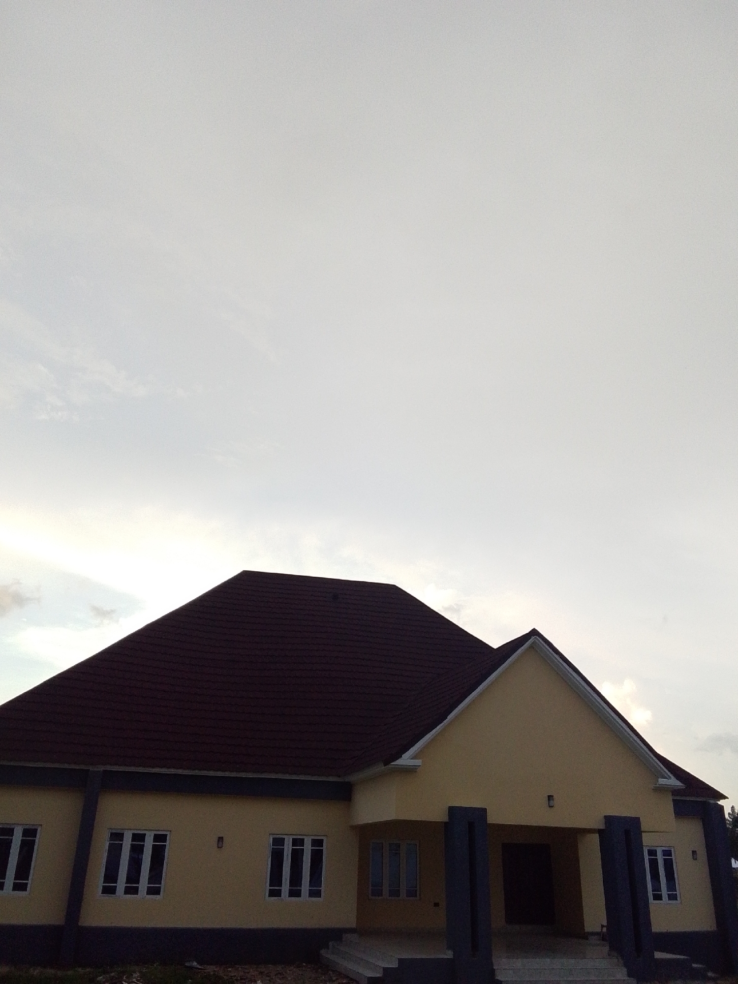 The image shows a yellow building with a dark red tiled roof, located in Serti, Nigeria. The building has multiple windows with white frames and the facade is accented with dark grey vertical columns. There are steps leading up to the entrance, which is partially obscured by the columns. The sky is overcast with light grey clouds. The ground in front of the building appears to be unpaved.