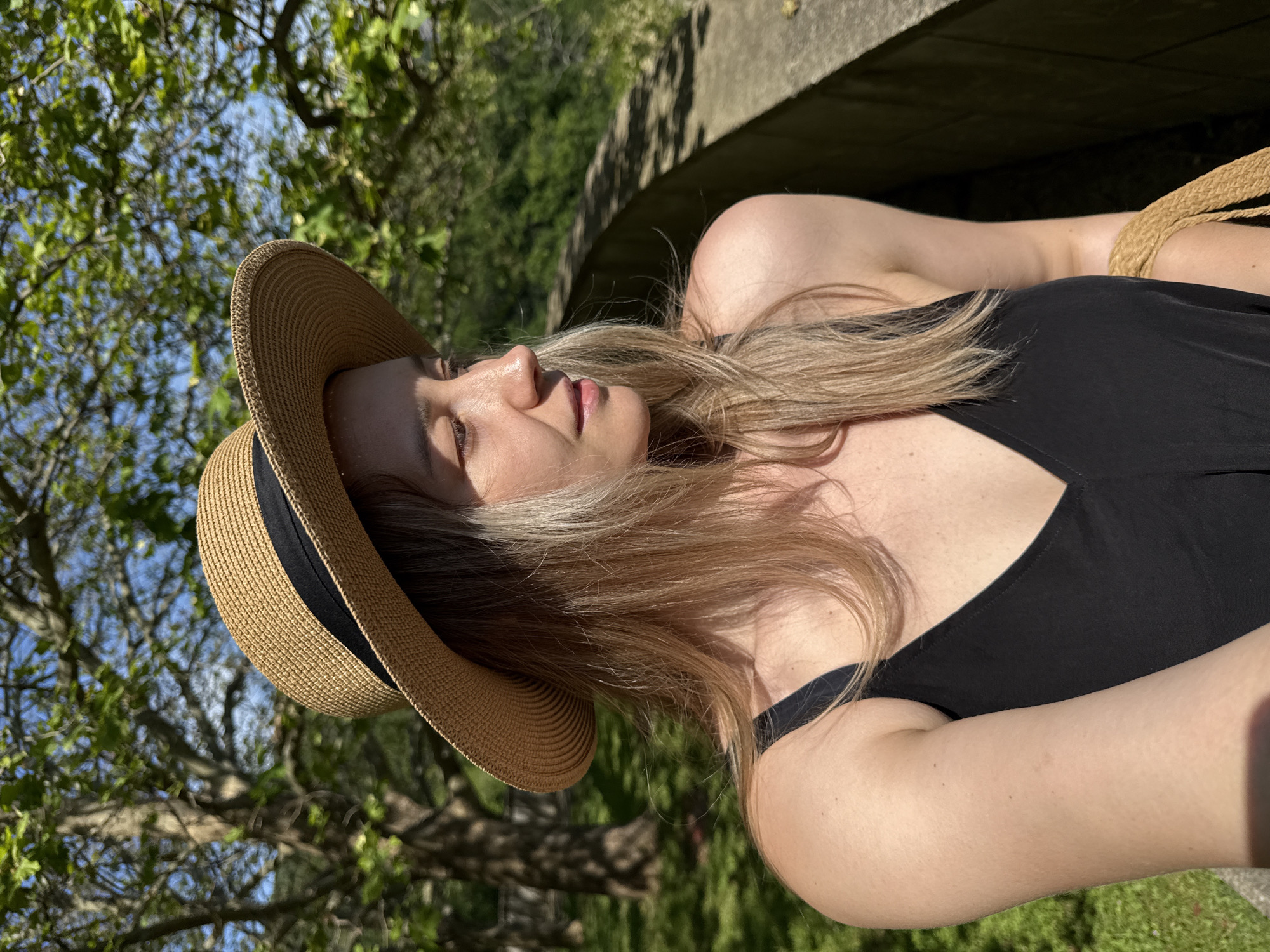 The image shows a woman wearing a straw hat and a black dress, standing outdoors in what appears to be a park or garden. She is looking to her right, with a soft smile on her face. The background features trees with green leaves and a blue sky. A stone wall runs along the right side of the image. The lighting suggests it is daytime, possibly late afternoon, with sunlight casting shadows. The woman is holding a straw bag on her right side, only a portion of which is visible. The scene evokes a relaxed and pleasant atmosphere. The provided crops offer close-up views of the woman's face, hair, shoulder, and the surrounding environment, highlighting details like the texture of her hat, the sunlight on her skin, and the foliage.