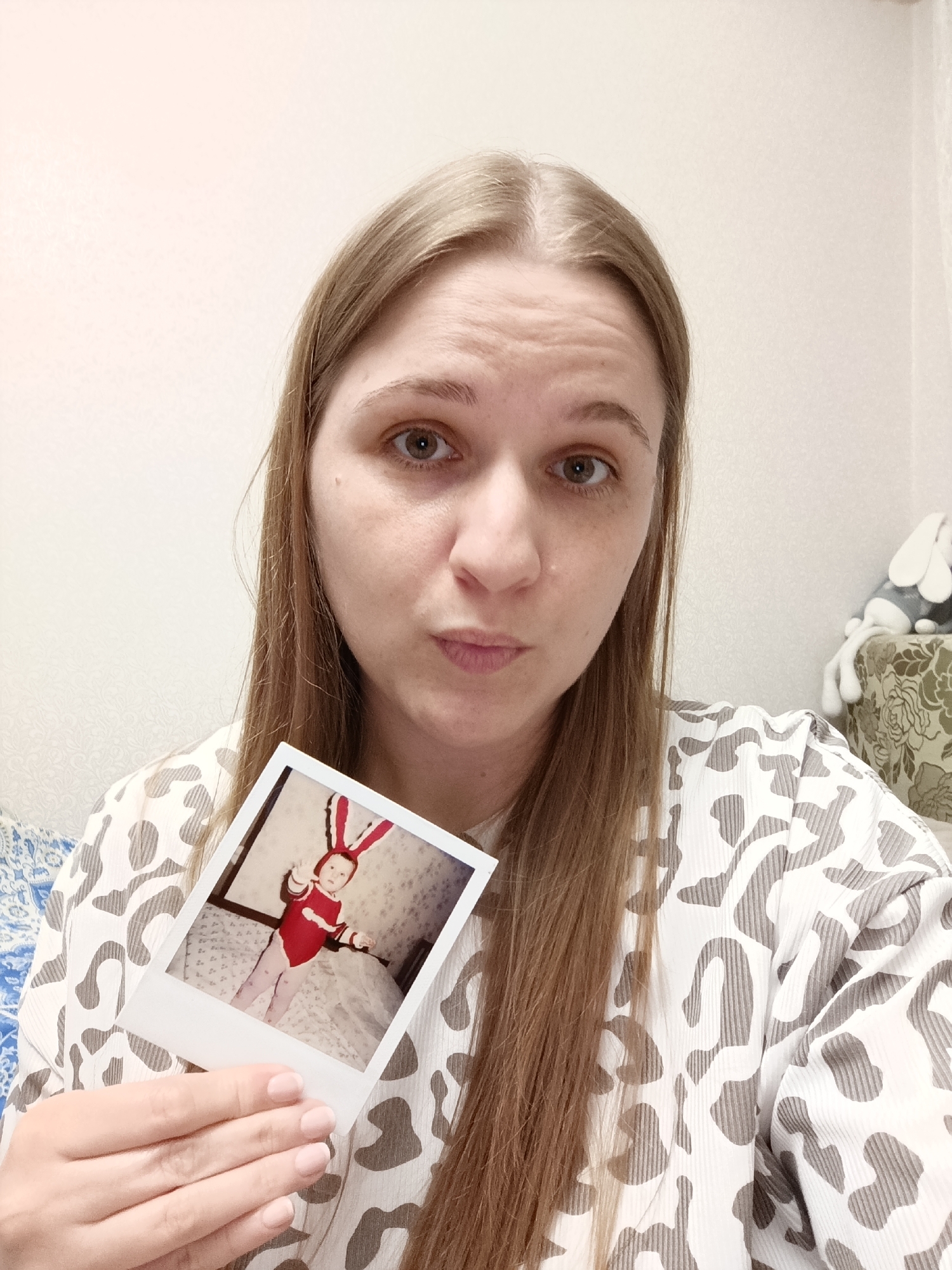A woman with long blonde hair and light eyes holds a Polaroid photo in her left hand. The photo shows a young child in a red bunny costume with bunny ears, standing on a bed. The woman is wearing a white and gray patterned shirt. The background appears to be a room with a white wall. The location is Unknown, Unknown. A stuffed animal is partially visible on a surface behind the woman. The woman has a slightly puckered expression.