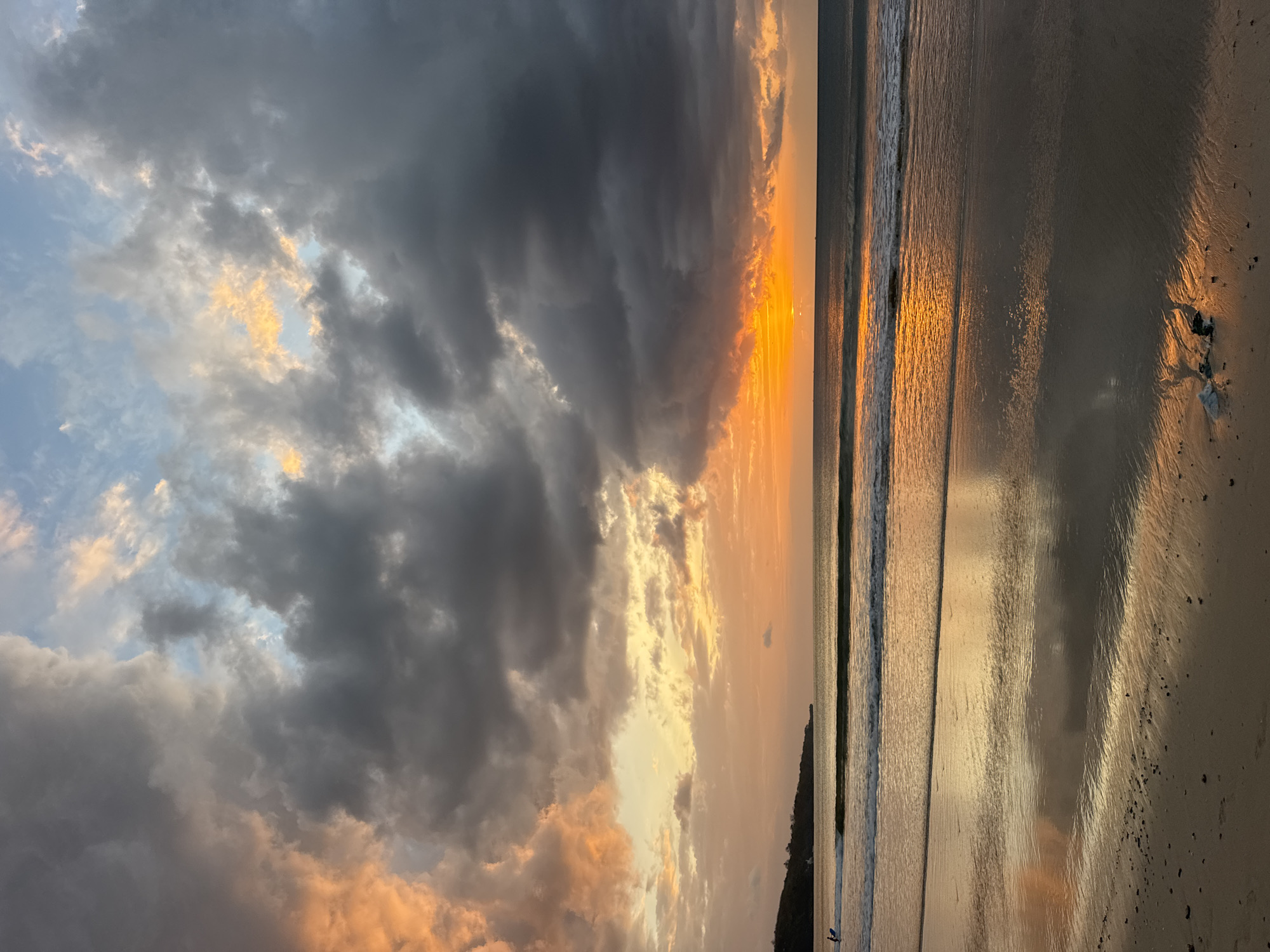 The image depicts a dramatic sunset over the ocean in Kuta Selatan, Indonesia. Dark, heavy clouds are illuminated from behind by the setting sun, creating streaks of fiery orange and soft pink across the sky. The ocean reflects the vibrant colors of the sunset, with gentle waves lapping at the wet sand of the beach. In the distance, a headland with vegetation juts out into the sea. A lone figure on a surfboard can be seen in the water, silhouetted against the bright horizon. The foreground of the beach is wet and sandy, with some small pebbles and debris visible.