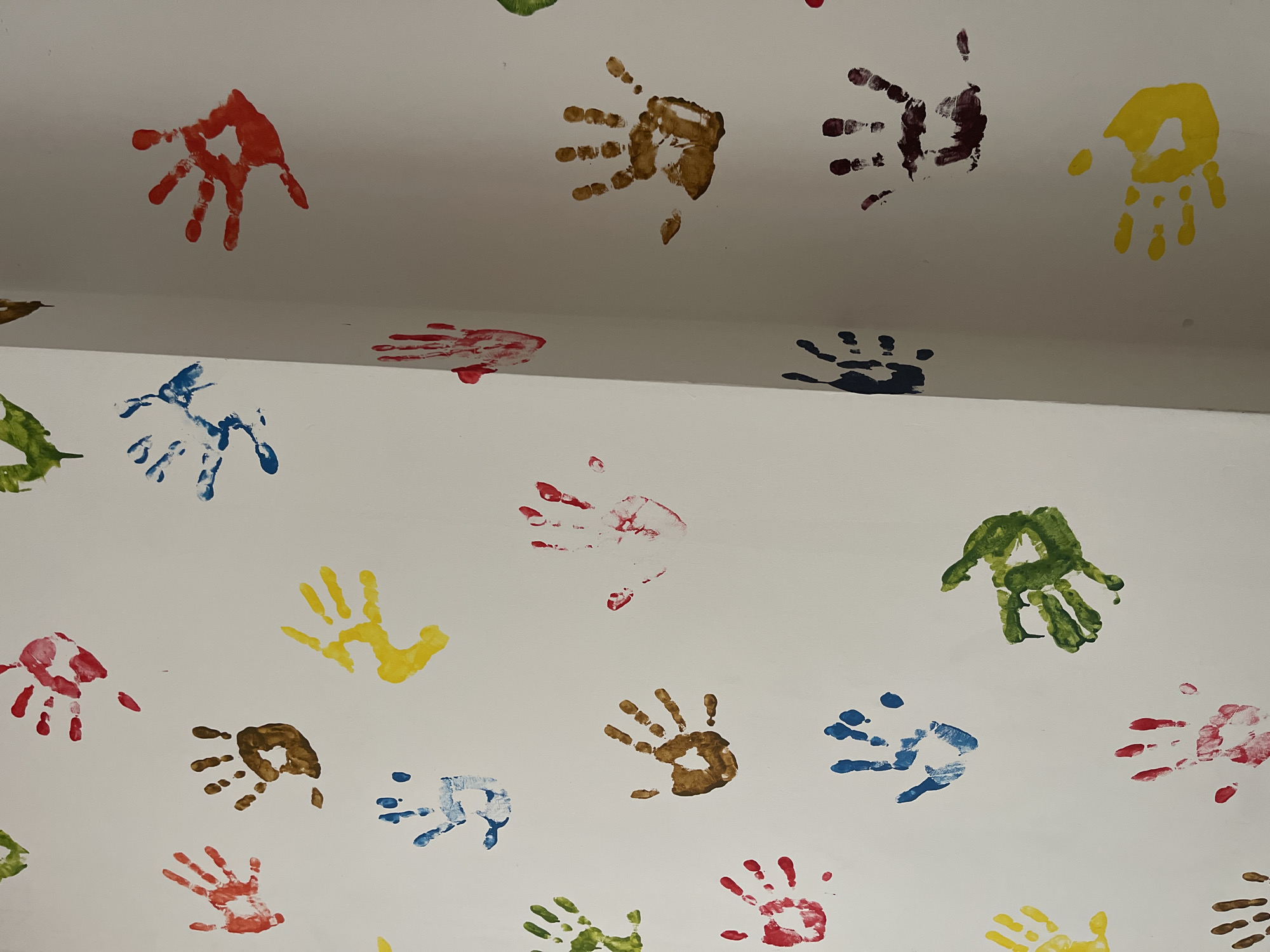 This is a close-up, eye-level, vertical, medium shot of a white wall adorned with colorful handprints. The handprints are varied in color, including red, brown, yellow, blue, green, and dark purple. They are scattered across the wall in a somewhat random pattern. Some handprints appear to be from children, while others might be from adults, judging by their size. The lighting is bright and even, indicating an indoor setting, possibly a child's play area, a classroom, or a decorated hallway. The focus is sharp on the handprints, with the wall texture slightly visible. The image appears to be captured in an unknown location in Unknown, Unknown.