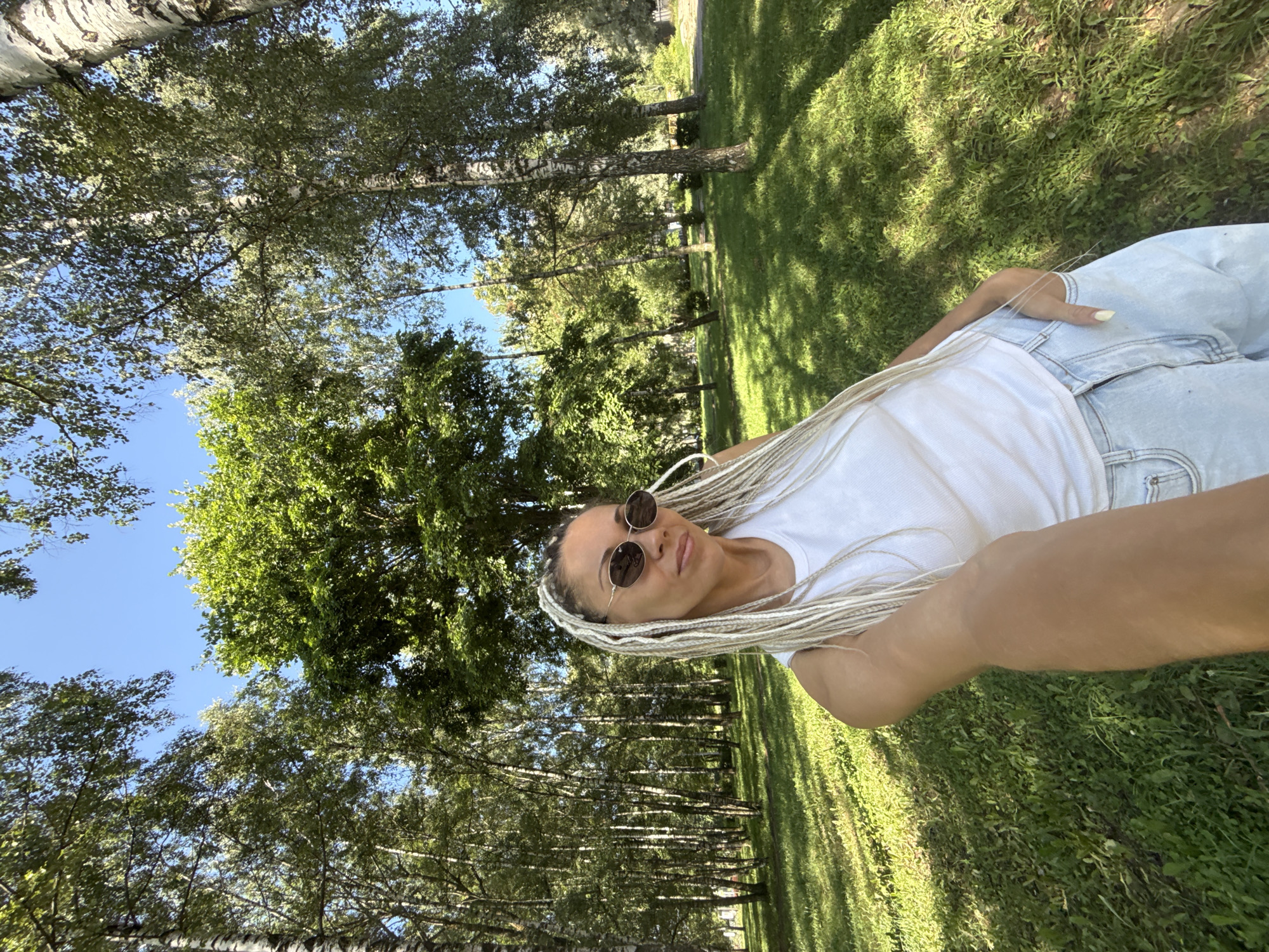 In Rusanovo, Russia, a woman with blonde braids is taking a selfie in a park. She is wearing a white tank top and light-wash jeans, with sunglasses on. The woman's right hand is in her jeans pocket. The background is filled with trees and lush green grass, with dappled sunlight filtering through the leaves. The sky is clear and blue.