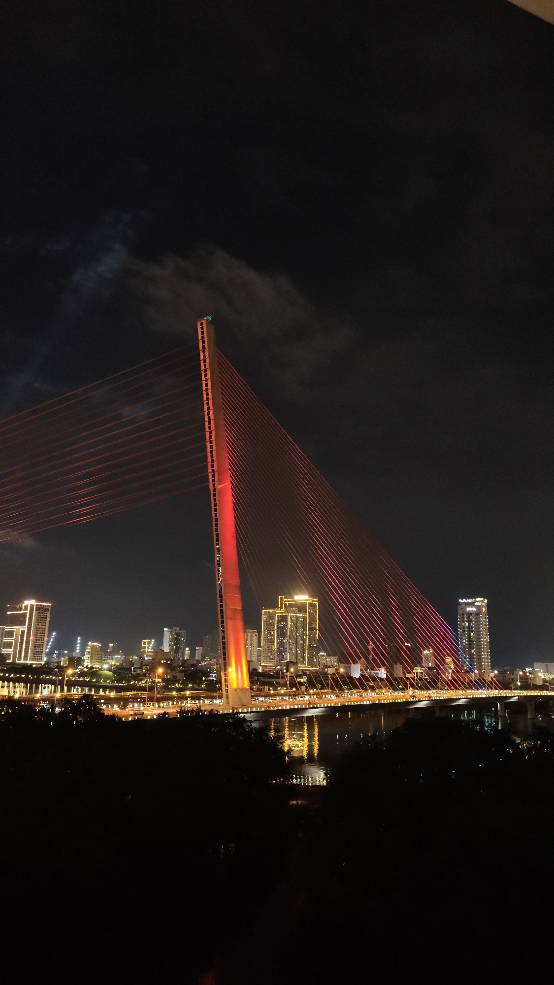 The image shows a bridge at night in Đà Nẵng, Vietnam. The bridge is a cable-stayed bridge with a tall, orange pylon. The cables are illuminated in red. In the background is a city skyline with various illuminated buildings. The foreground shows dark trees or foliage. The water reflects the lights from the bridge and the city. The sky is dark with some clouds.