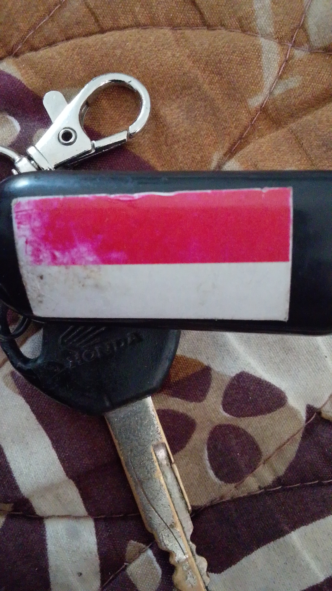 A close-up, overhead shot features a keychain with a Honda logo and a motorcycle key resting on a patterned fabric. The keychain's metal clip is visible at the top, attached to a black cylindrical object. A rectangular sticker with red and white stripes, resembling the Indonesian flag, is affixed to the cylinder. The motorcycle key, made of metal, is positioned horizontally across the fabric. The fabric has a repeating pattern of brown and beige shapes. There is no discernible information about a specific location in Cintakarya, Indonesia.
