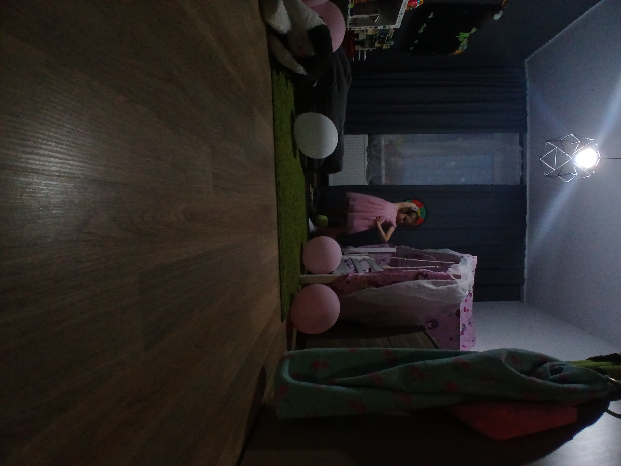 The image depicts a dimly lit room, likely a child's bedroom. In the center, a young girl in a pink dress and a festive hat stands with her hands on her hips. Several large, round pink balloons are scattered on the floor, along with a white balloon. To the left, a stuffed animal rests on a dark surface, possibly a blanket or pillow. In the background, dark curtains cover a window, and a child's play tent with a pink and white pattern is visible. A modern geometric light fixture hangs from the ceiling, and a patterned textile is draped over a hook on the right side of the frame. The floor is made of dark wood laminate. There are no identifiable landmarks or context suggesting the location is Yekaterinburg, Russia.
