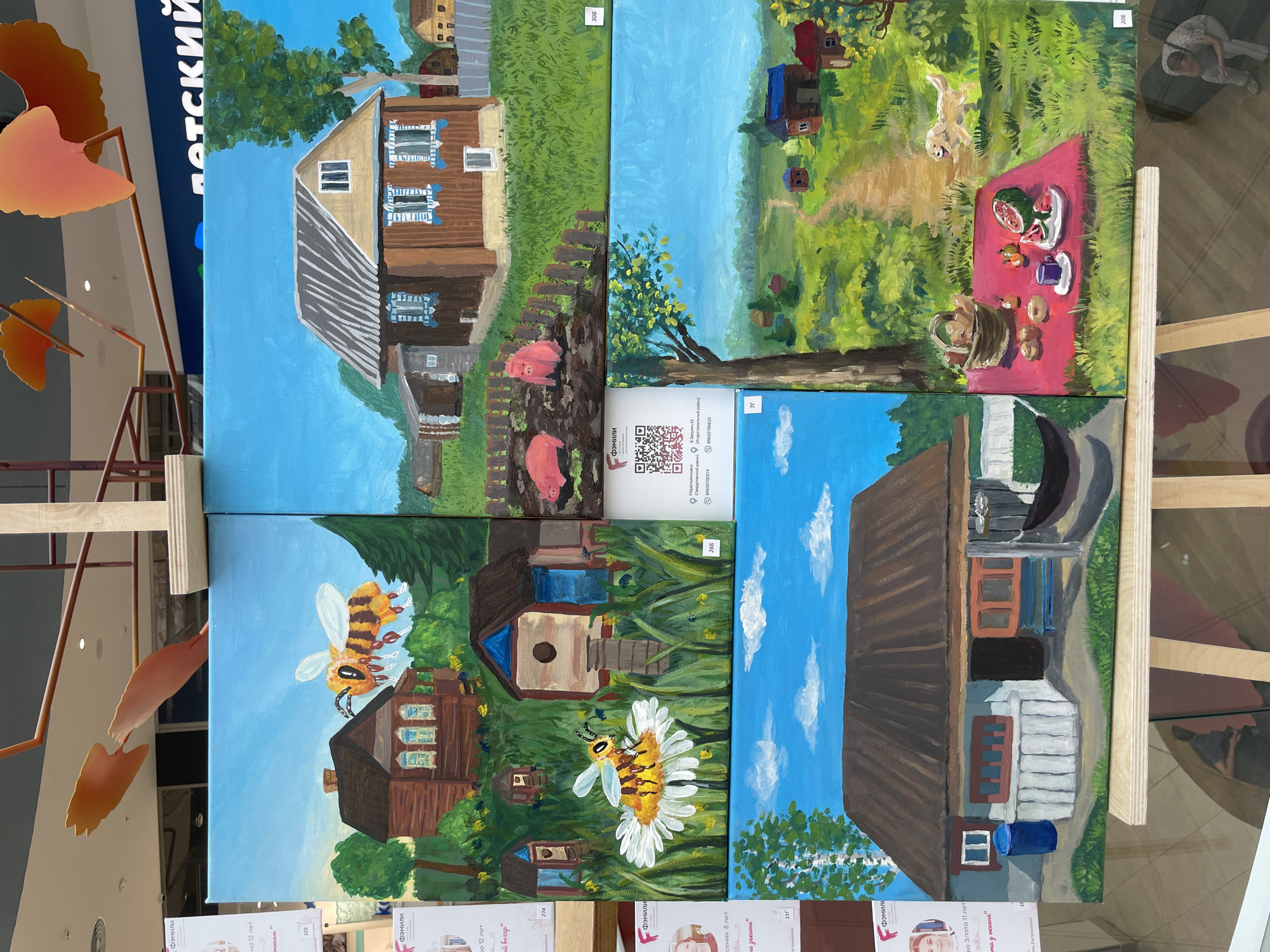 This media file displays a collection of four acrylic paintings mounted on easels. The paintings appear to be the work of children or young artists, given the "Фэмили" (Family) branding visible on posters beneath some of the artworks, suggesting a family art program or exhibition. The paintings depict rural or suburban scenes with a naive, charming style.
The top-left painting shows a wooden house with blue shutters and a thatched roof, a wooden fence, and two pink pigs in a yard. The top-right painting features a sunny, green meadow with a path leading to a small cluster of houses. A dog is captured mid-leap, and a picnic blanket is spread out with watermelon, fruit, and a basket.
The bottom-left painting is a close-up of a bee on a white daisy, with several small beehives and a larger wooden house in the background, all set against a bright blue sky. The bottom-right painting depicts a simple wooden house with red and blue window frames, a dark roof, and a blue barrel at its base, also under a bright blue sky with white clouds.
The overall mood of the paintings is cheerful and idyllic, suggesting summer days and rural life. The bright colors and simplified forms evoke a sense of innocence and joy. The location context of Perm, Russia, is hinted at by the Cyrillic text on the supporting posters, which include addresses like "Р. Крестьянская, 6" and "В.Засулич 42," along with phone numbers. The phrase "детский" (children's) is also partially visible on a blue banner in the background.
