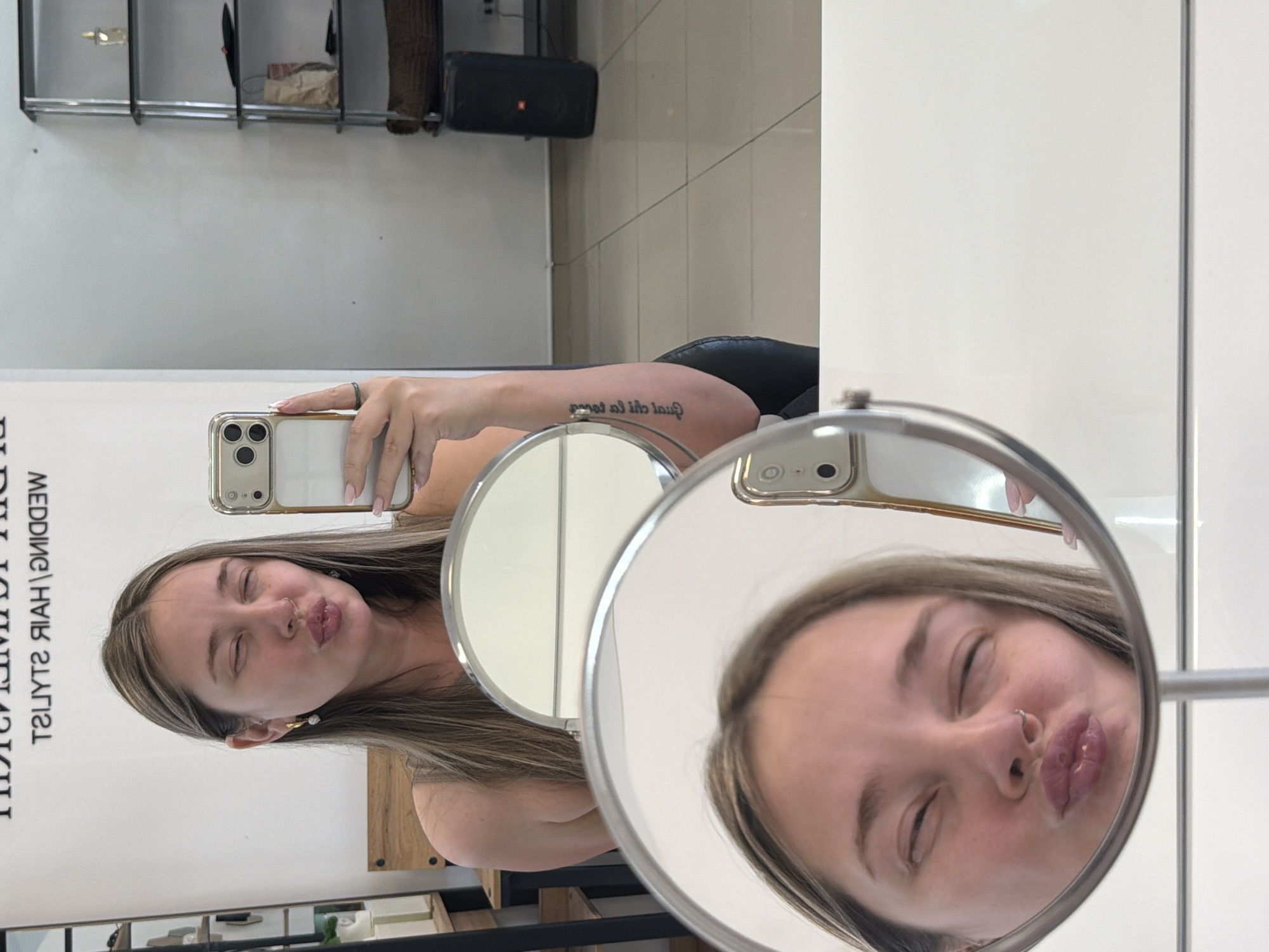 A young woman is taking a selfie using an iPhone 13 or similar model. She is visible directly and through a small circular mirror. The woman has light brown hair with blonde highlights and appears to be puckering her lips in both reflections. A faint tattoo is visible on her left forearm. The photo seems to be taken in what appears to be a beauty salon or similar establishment. There is shelving behind her and the text "HAIR STYLIST" appears, although the rest is cut off. The location is likely Chaikovsky, Russia.