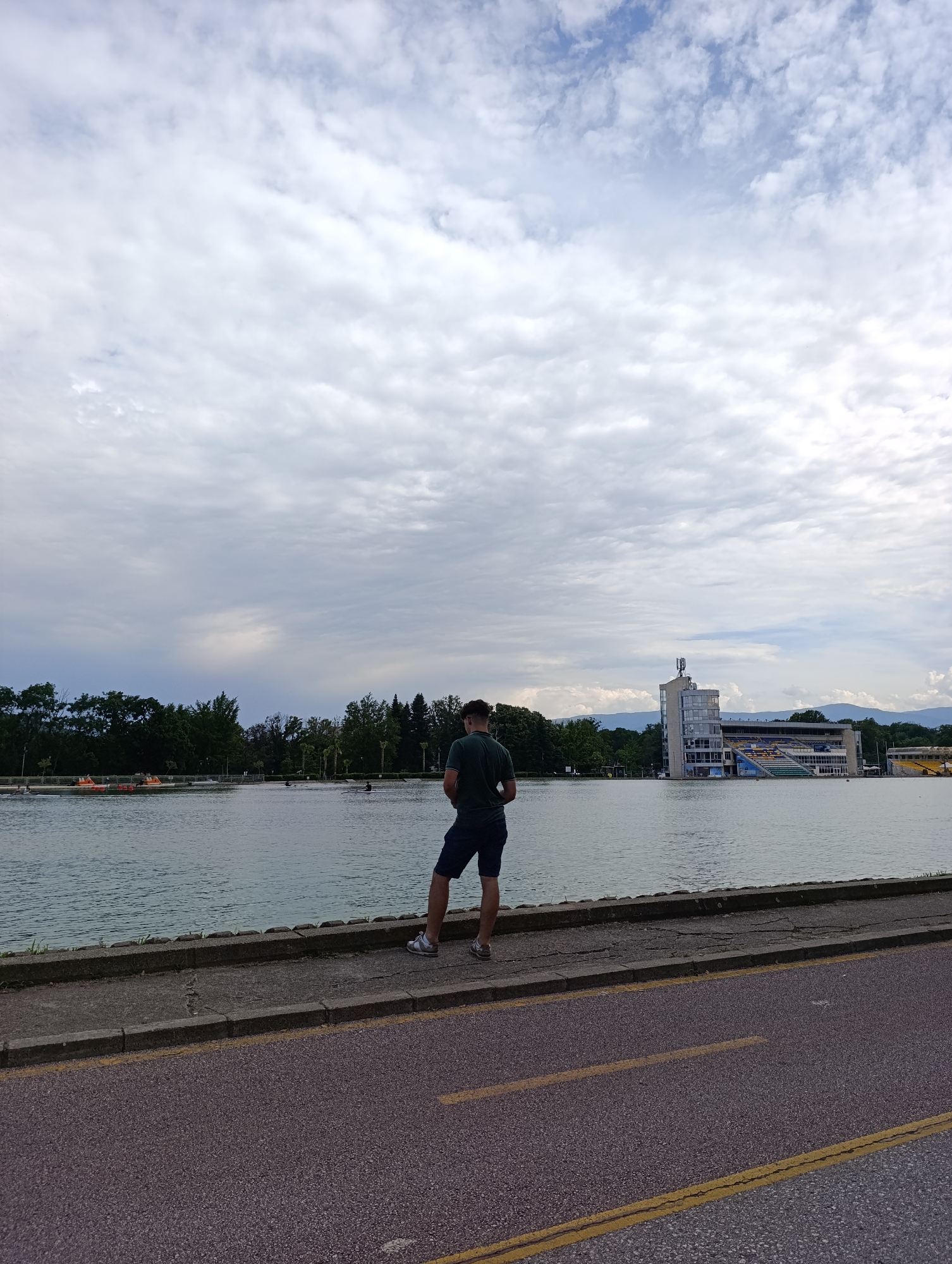 The image presents a wide outdoor view of a person observing a large body of water under an overcast sky. In the foreground, a person is seen from behind, standing on a paved walkway at the edge of the water. They are wearing a dark green t-shirt, dark shorts, and sneakers, looking out over the water.
The body of water, resembling a rowing canal or lake, stretches across the mid-ground. The far bank is lined with a dense array of green trees. To the right, a prominent multi-story concrete building with a tall tower and grandstands featuring visible yellow and blue seating stands by the water, indicative of a sports complex or regatta venue. In the hazy distance beyond the trees and buildings, faint outlines of mountains or hills are visible.
Activity on the water includes several small boats. Towards the left, bright orange or yellow pedal boats are near the bank, while further out, individuals in dark, narrow rowing boats can be seen engaged in rowing.
The immediate foreground features a paved path with visible cracks, adjacent to a curb. Beyond the curb, a grey asphalt road with a double yellow center line runs parallel to the water. The sky is largely covered with a mix of white and grey clouds, creating an overcast but bright daytime atmosphere. This scene is consistent with the Regatta Venue in Plovdiv, Bulgaria. No explicit text is visible in the image.
