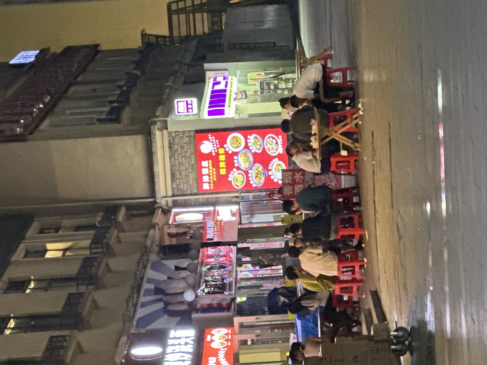 The image depicts a nighttime street scene in the city of Chongqing High-tech Zone, China. A group of people are gathered around several tables outdoors, seated on red stools. Some individuals appear to be eating, with plates and drinks visible on the tables. The tables are small and appear to be made of wood. In the background, there are buildings with illuminated signs and windows, suggesting a commercial area. The street surface is wet, reflecting the ambient light. The overall impression is of a casual dining or social gathering taking place after dark.