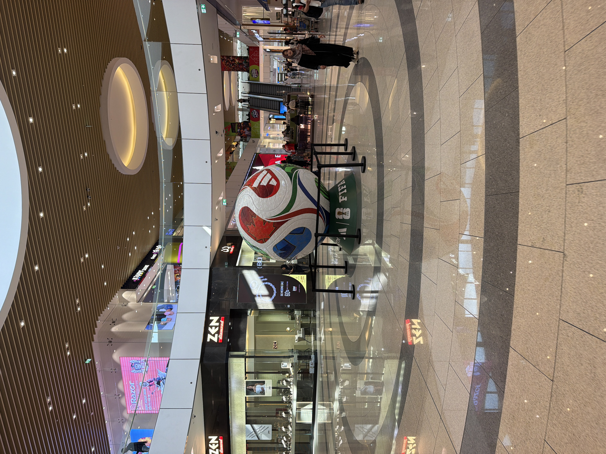 This media file captures a wide shot of an indoor shopping mall, likely in a temperate climate given the presence of shoppers. The scene is brightly lit, suggesting daytime. The primary focus is a large, stylized soccer ball displayed in the center of a walkway. The ball is red, white, and blue with intricate paneling, and it rests on a green platform with the FIFA logo clearly visible.
Several people are scattered throughout the mall. In the background, there's a woman with a headscarf wearing a long black coat, walking away from the camera. Further in the distance, more individuals are seen browsing shops, ascending escalators, and congregating at seating areas. The mall features a modern architectural design with high ceilings, polished tiled floors, and curved white structures with embedded lighting. Shopfronts are visible on either side of the walkway, some with illuminated signs and displays. One sign reads "ZEN diamond" and another advertises a "BIG WEDDING SALE 60%". Another digital display shows "Razor" and some gaming-related imagery. The overall atmosphere appears to be calm and casual, indicative of a typical shopping day. The orientation of the image is such that the bottom of the frame is to the left, and the top is to the right, with the main subjects oriented vertically.