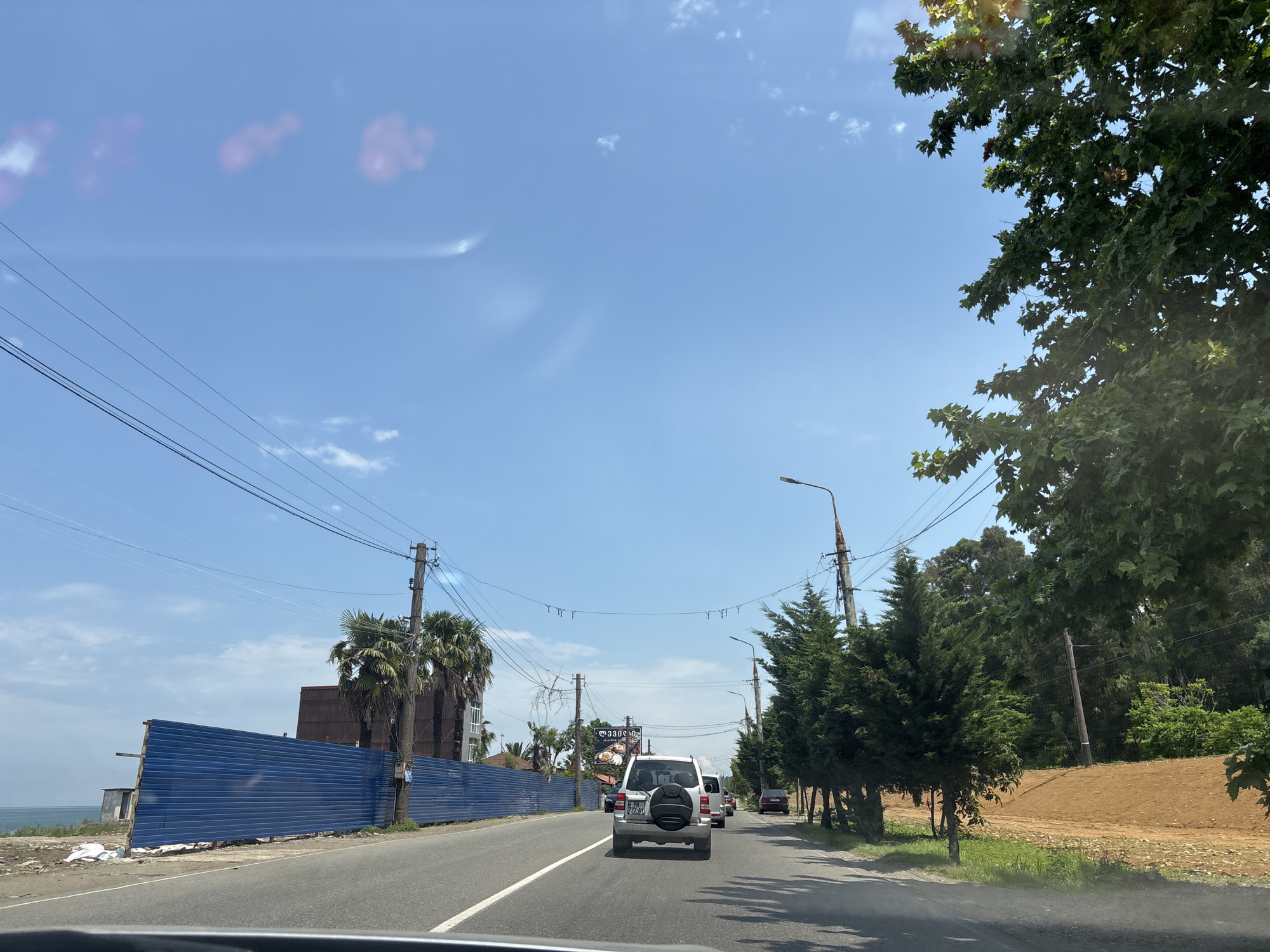 The image depicts a street scene in Batumi, Georgia, on a clear, sunny day. The perspective is from inside a vehicle, looking forward along a paved road. The road is two lanes wide with a white dividing line and shoulders on both sides.
On the left side of the road, a long, blue corrugated metal fence runs along the scene, likely indicating a construction site. Beyond the fence, a glimpse of the sea is visible. Further down the road, several tall palm trees stand, and a building with a reddish-brown roof is partially visible. Overhead, multiple power lines crisscross the sky, supported by utility poles.
On the right side of the road, a row of evergreen trees lines the shoulder, followed by a mound of orange-red soil. Beyond the trees, dense green foliage suggests a wooded area. Several streetlights are positioned along the road.
Several vehicles are on the road, including a white SUV in the foreground, followed by other cars. The SUV has a spare tire mounted on its rear. The sky is a vibrant blue with scattered wispy white clouds. The overall impression is a coastal urban or suburban environment with ongoing development.
