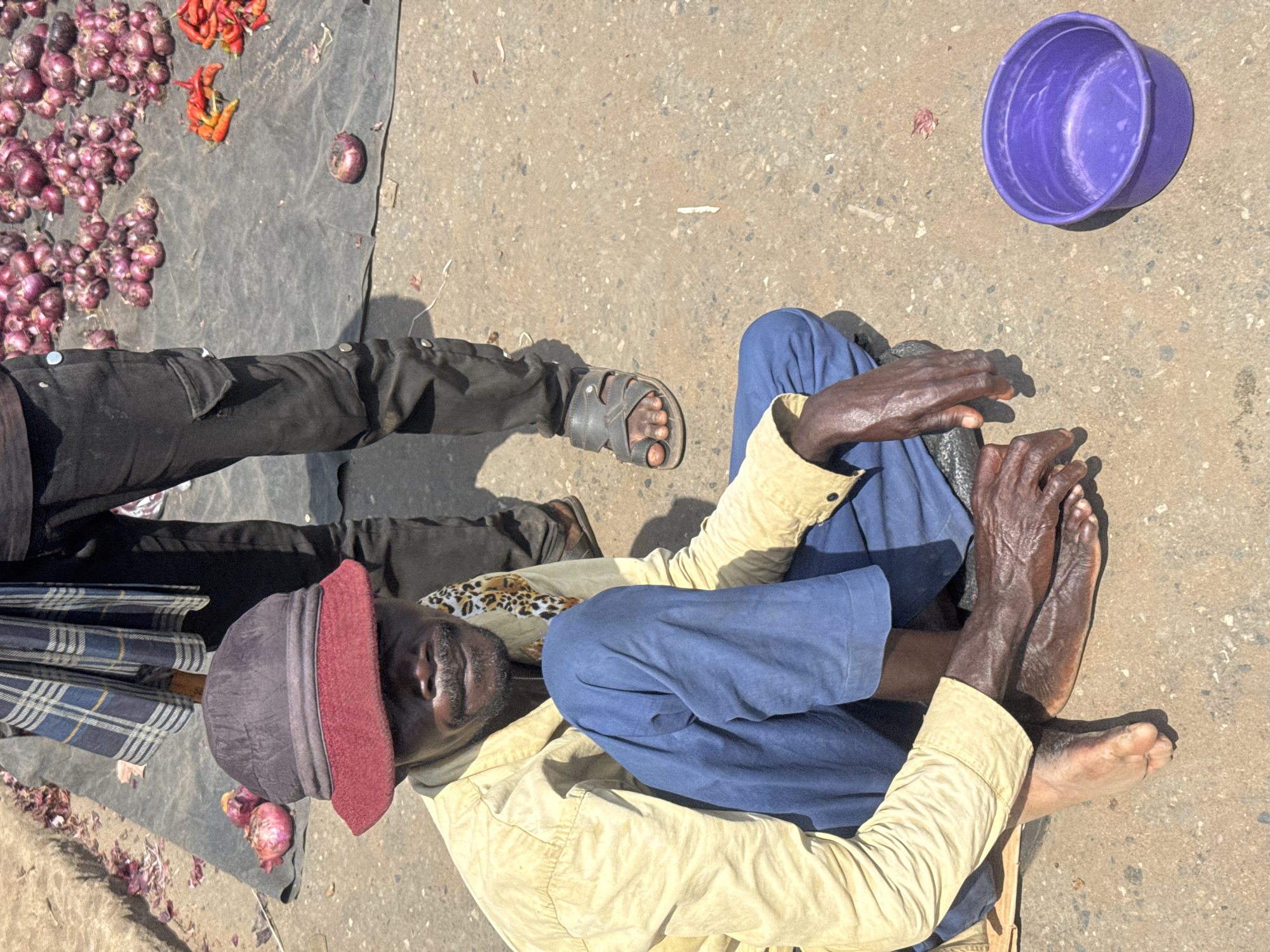 This overhead shot depicts an outdoor market scene in Onitsha, Nigeria, under bright, direct sunlight, casting strong shadows.
The central figure is an older man seated on the ground with his legs bent. He has dark skin, and visible wrinkles on his hands and feet, indicating age. He is barefoot and wears a light-colored, long-sleeved shirt (khaki or tan), blue pants, and a dark cap with a red brim. His expression appears neutral.
To his left, the lower legs and feet of another individual are visible, standing. This person wears black pants and grey sandals.
On the ground, a black tarp is laid out, displaying produce for sale. Large piles of small red onions dominate the left side of the tarp, alongside a smaller cluster of red and yellow chili peppers. A single purple onion rests alone on the tarp towards the right. Nearby, a plain purple plastic bowl sits on the concrete. A folded plaid umbrella with a blue, black, and white pattern is positioned next to the seated man, partially on the tarp.
The setting appears to be a bustling street or market area, characterized by the informal display of goods directly on the ground. No visible text is present in the image.