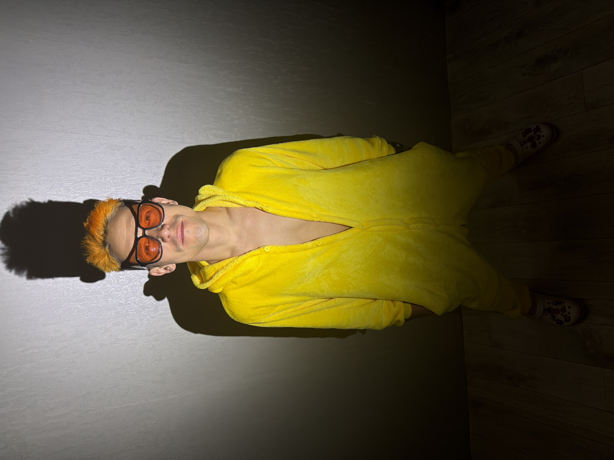 A man is lying on his side. He is wearing a yellow, fleece onesie, orange tinted glasses and he has orange hair. The setting appears to be indoors on a light gray tiled floor, with a dark wooden floor behind him. The lighting is dim, casting a large shadow to his left. There is no evidence to verify the location.