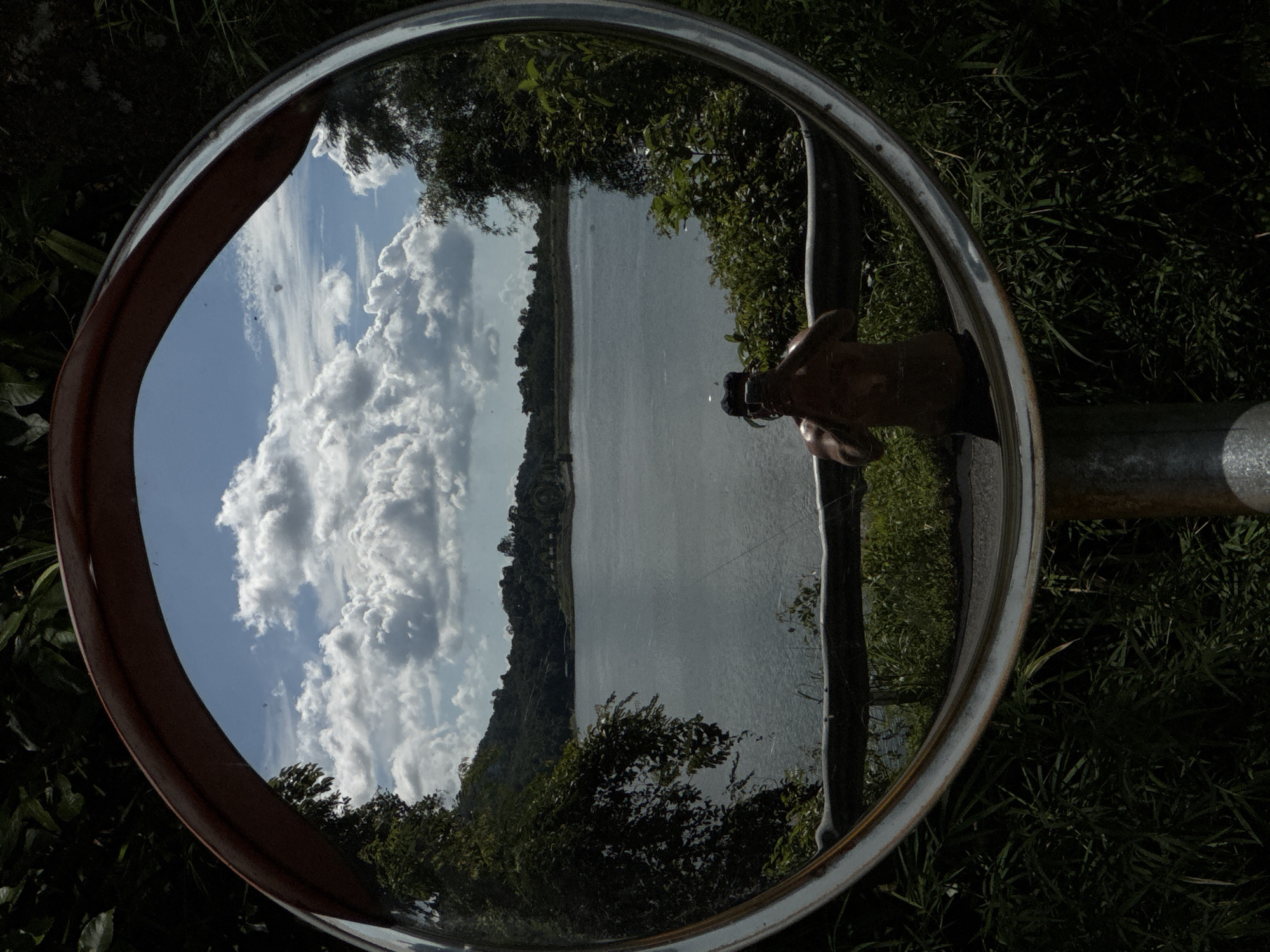 A convex mirror is mounted on a metal pole, reflecting a scenic view of a large body of water, a forested hillside, and a partly cloudy sky. In the foreground, a person is visible in the reflection, holding a camera and taking a selfie. The person appears to be shirtless and is positioned in front of a railing, with lush green foliage surrounding the mirror and extending into the background. The overall scene suggests an outdoor location, possibly a reservoir or lake, in Kathu, Thailand, given the lush vegetation and the bright, diffused sunlight indicative of a tropical climate.