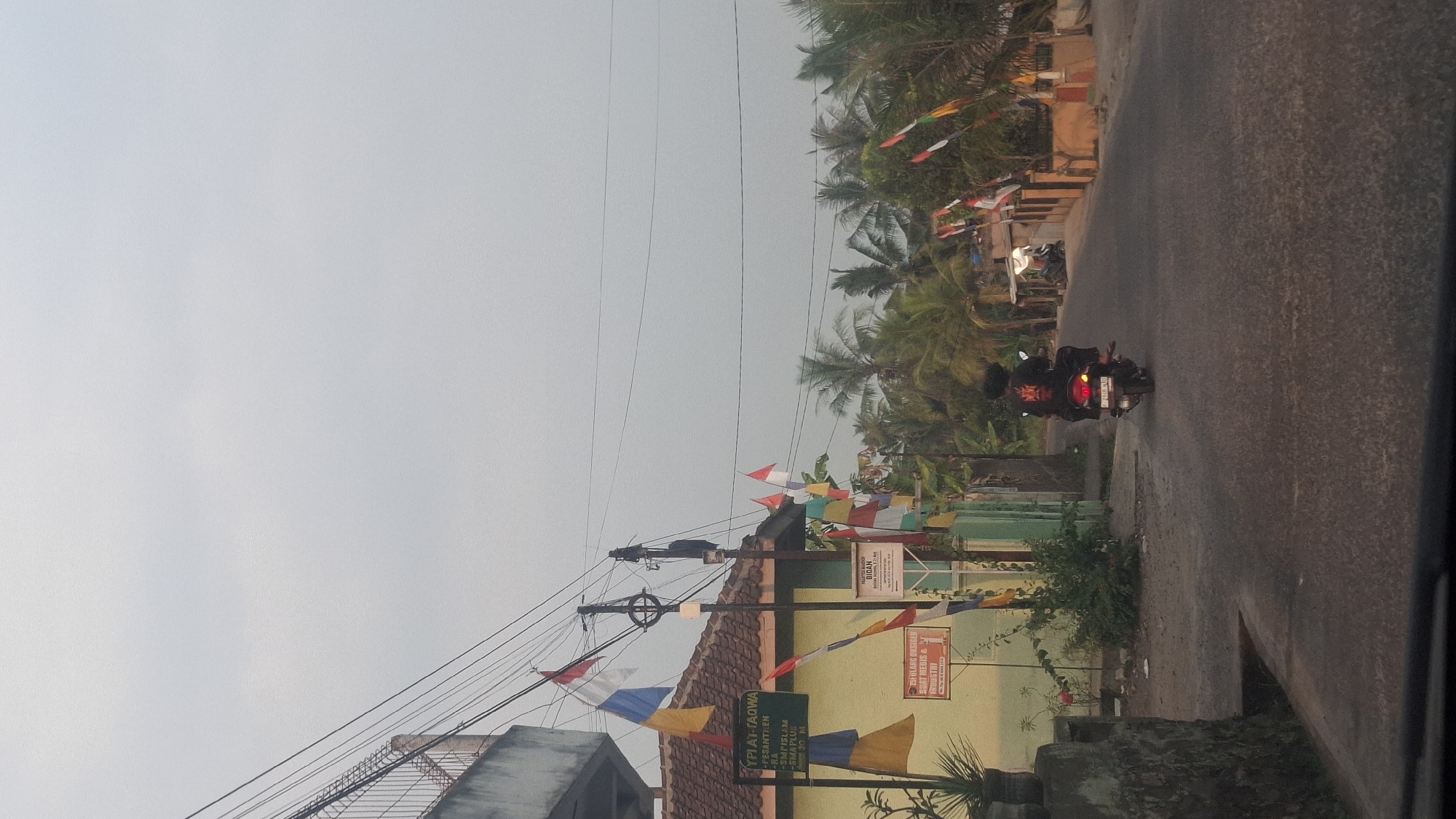 The image depicts a street scene in Cibinong Hilir, Indonesia, characterized by a light overcast sky. In the foreground, the road surface is visible, with a person riding a motorcycle away from the camera. The motorcycle is black and has a license plate that is partially obscured. The rider is wearing a black shirt with a red emblem on the back.
To the left of the road, there is a building with a yellow facade and a tiled roof. A sign on the building reads "YPI AT-TAQWA" followed by what appears to be "PESANTREN" and then "SMA PLUS". Numerous small, colorful flags, predominantly red and white, are strung across wires above the building and the street, suggesting a festive or patriotic occasion. Further to the left, a stone wall or structure is present.
On the right side of the road, a line of palm trees and other vegetation is visible. Behind the trees, there are several houses with various roof colors, and more flags are displayed. A smaller sign with text is mounted on a pole near the building on the left. The overall impression is that of a residential street in a tropical or subtropical climate.