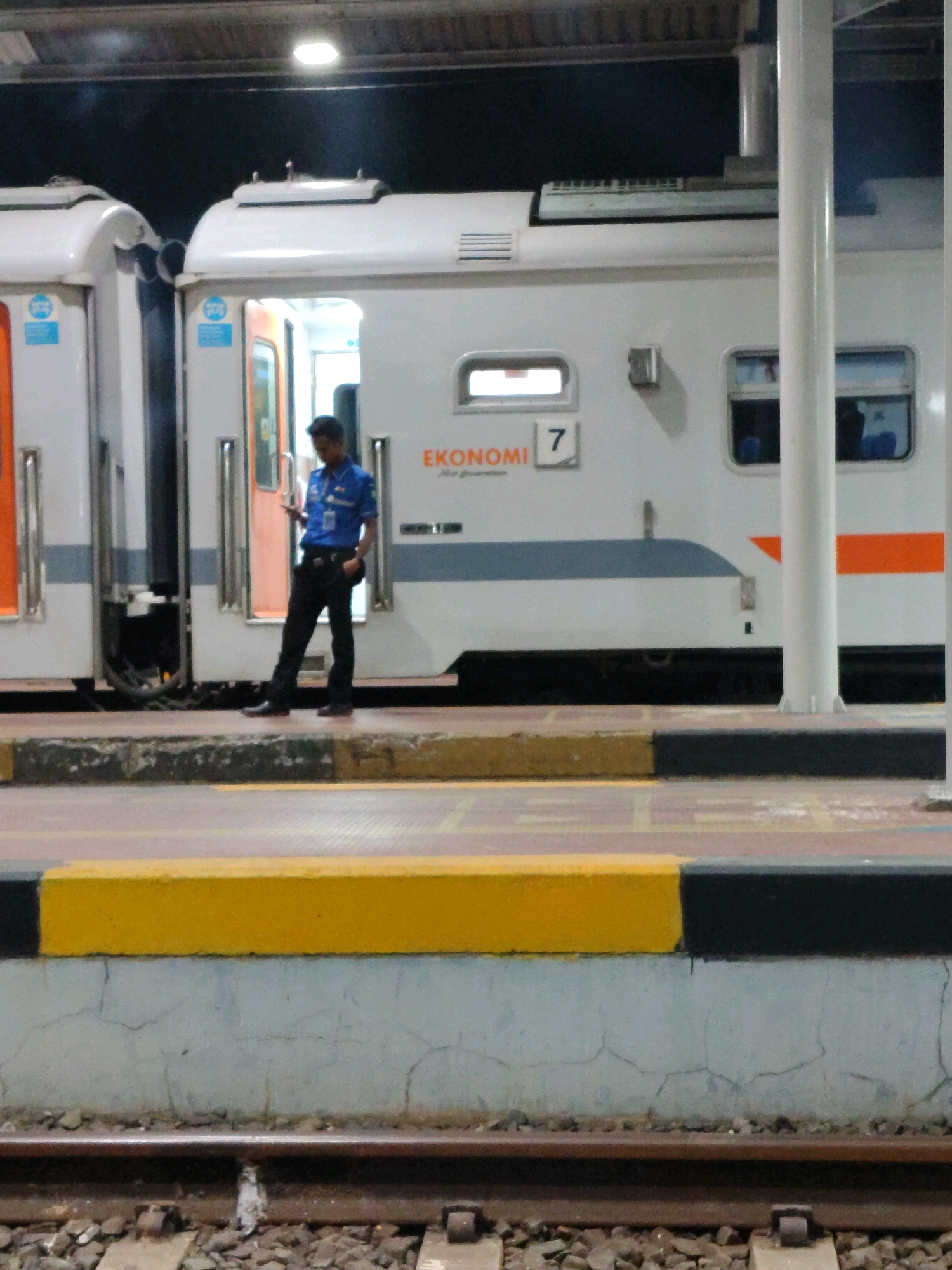 An outdoor, eye-level medium shot captures a man in a blue uniform with black pants standing on a train platform in Jatibarang, Indonesia. He is holding a mobile phone in his right hand, looking down at it. To his left, a white train car with orange and gray trim is partially visible. The train has a door open, and the text "EKONOMI 7" is visible on its side. The platform is edged with a yellow and black stripe. In the foreground, train tracks are visible. The lighting suggests it is nighttime or dusk.