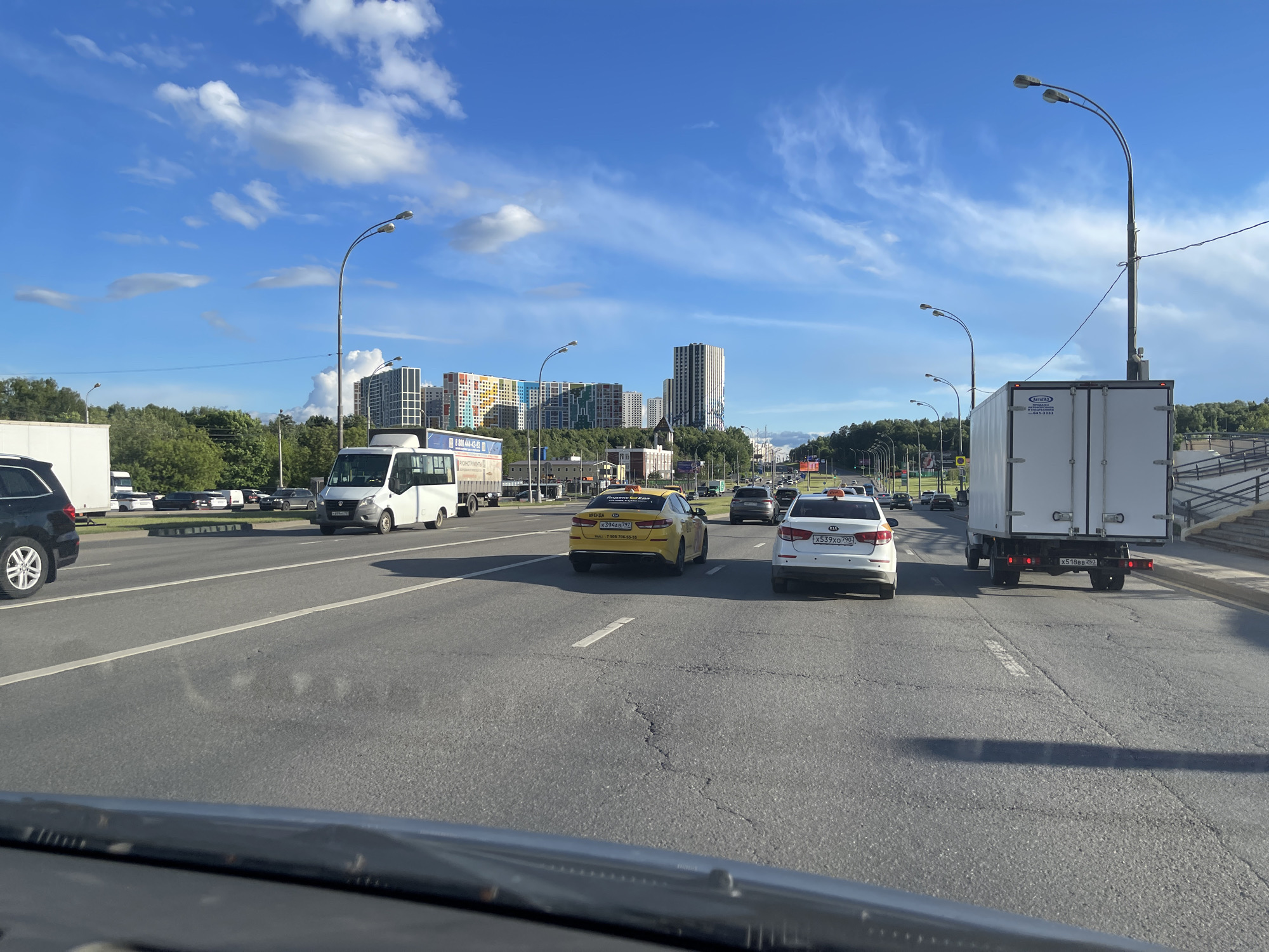 The image captures a multi-lane road in Moscow, Russia, on a sunny day with blue skies and scattered clouds. The view is from inside a vehicle, likely the driver's perspective. Several cars are present, including a yellow taxi, a white sedan, and a white truck. Buildings are visible in the background beyond a line of trees. Street lamps line the side of the road. The road shows signs of wear and tear.