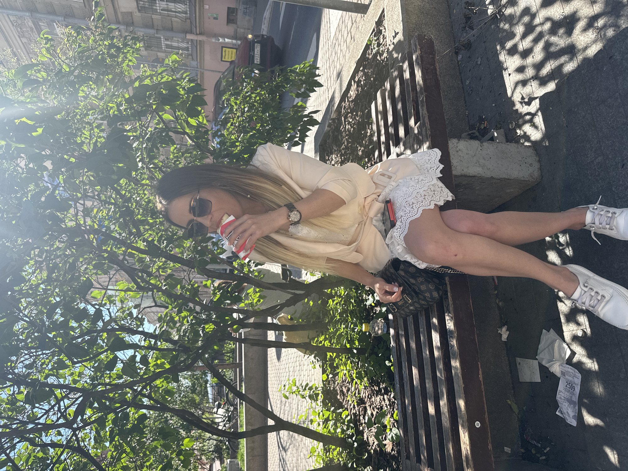 A woman is sitting on a park bench in Saint Petersburg, Russia, holding a disposable cup and drinking from it. She is wearing sunglasses, a light-colored dress with white lace trim, and white sneakers. A designer handbag is placed on the bench next to her. The scene is outdoors, with trees and dappled sunlight casting shadows on the ground. The weather appears to be sunny and pleasant, suggesting a daytime setting. There is some litter on the ground near the bench. A parked car and buildings are visible in the background. There is text on a yellow sign in the background which reads "PRODAЖA" (SALE). Another piece of paper on the ground has the number "2509" visible. The woman appears relaxed, enjoying a drink in a public outdoor space.