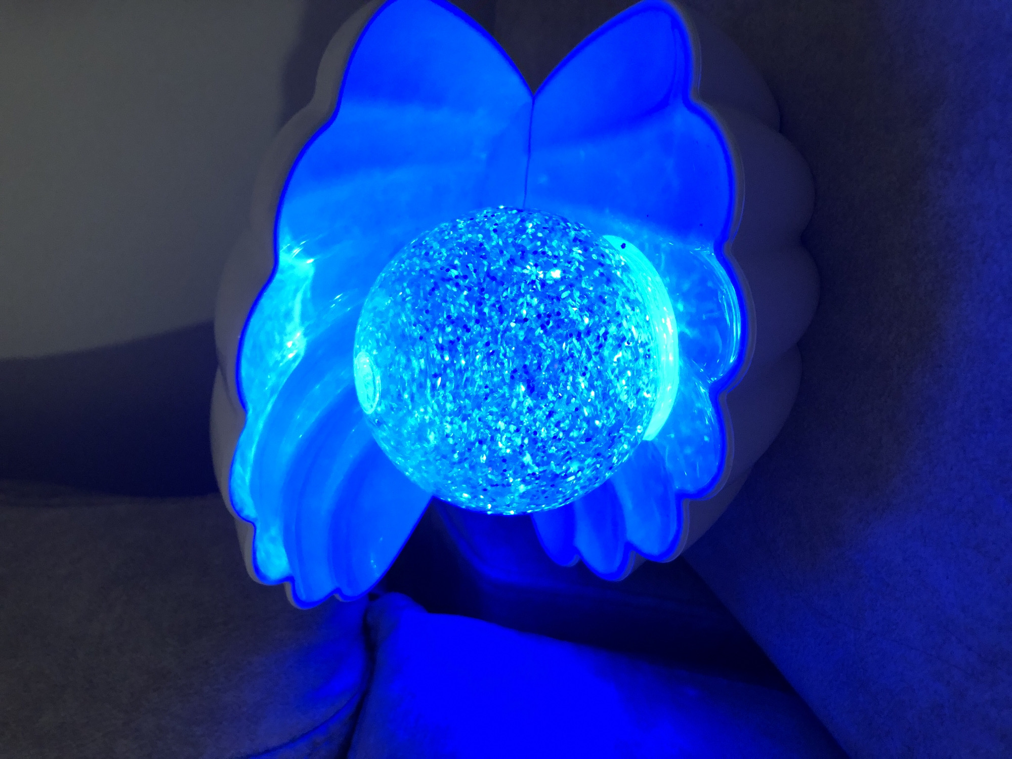This is a close-up, low-angle shot of a decorative object illuminated by bright blue light. The object appears to be a sphere covered in small, reflective particles, possibly glitter, giving it a sparkly appearance. This sphere is cradled within a white, petal-like structure that is also glowing with the same intense blue light. The petals have a scalloped edge and create a flower-like or winged silhouette around the central sphere.
The background is dark and appears to be soft, possibly fabric or upholstery, with areas of deep shadow and areas reflecting the blue light. The overall impression is that of a decorative light fixture or a fantastical object, possibly a part of a larger installation or a personal item. The lighting is dramatic, with strong contrasts between the illuminated areas and the shadows. The image is static, with no discernible activity or event taking place beyond the object itself being lit. There is no visible text, and cues about time of day, weather, or specific location within València are absent, though the electric lighting suggests an indoor setting or nighttime. The dominant colors are bright blue and white, with dark grays and blues in the background.