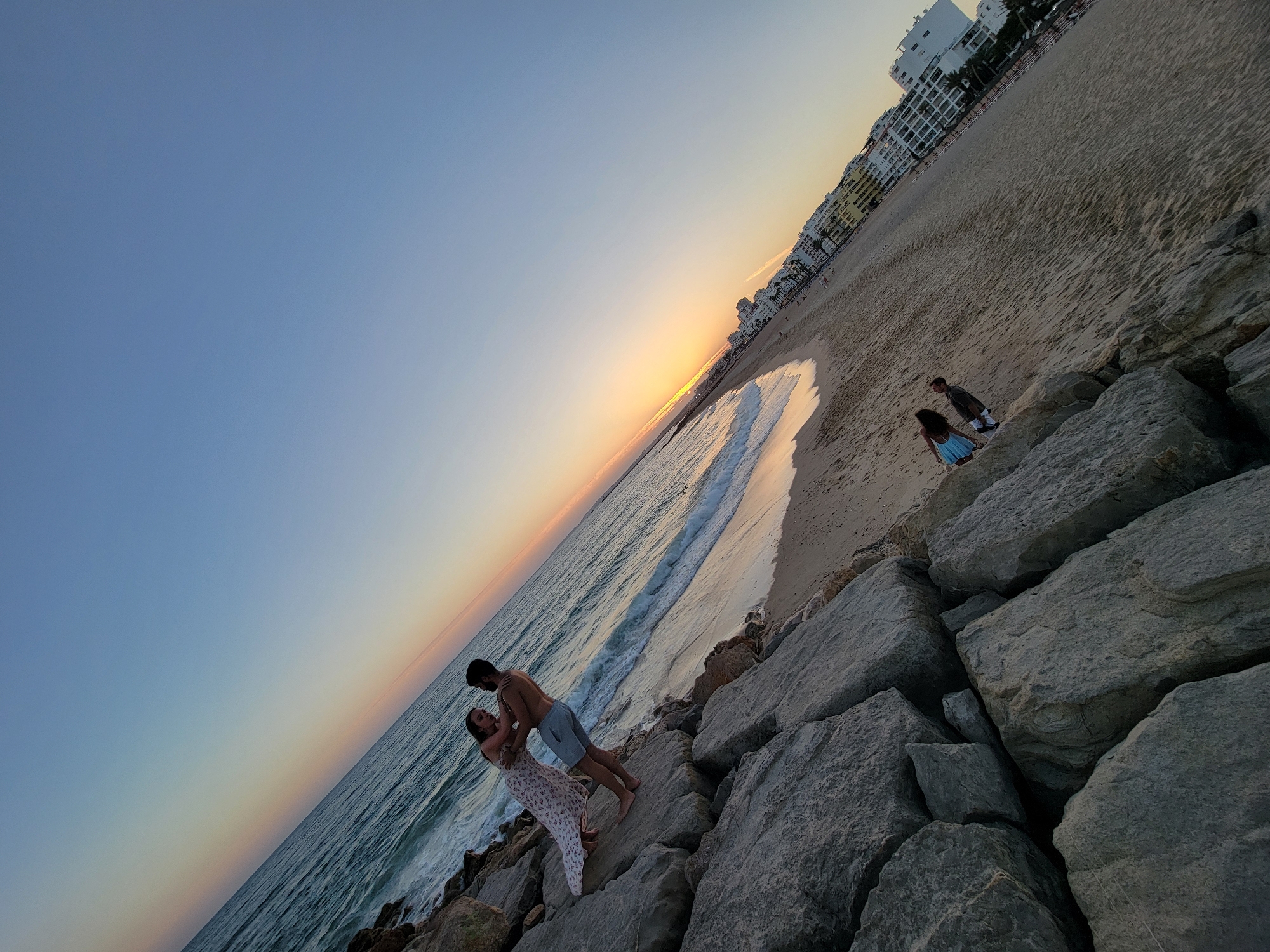 The image depicts a couple embracing on a rocky shoreline in Quarteira, Portugal, during sunset. The couple is positioned on large, grey rocks that extend into the ocean. The woman is wearing a white, floral-patterned dress, and the man is shirtless, wearing light-colored shorts. In the background, a sandy beach stretches along the coastline, lined with white buildings and palm trees, indicative of a resort town. The ocean water is calm with gentle waves lapping the shore. The sky displays a gradient of colors, transitioning from a pale blue at the top to warmer hues of orange and yellow near the horizon, characteristic of a sunset. The composition is angled, with the shoreline running diagonally across the frame. Further down the beach, other people can be seen, as well as a few scattered beach chairs and umbrellas. The overall scene conveys a serene and romantic atmosphere.