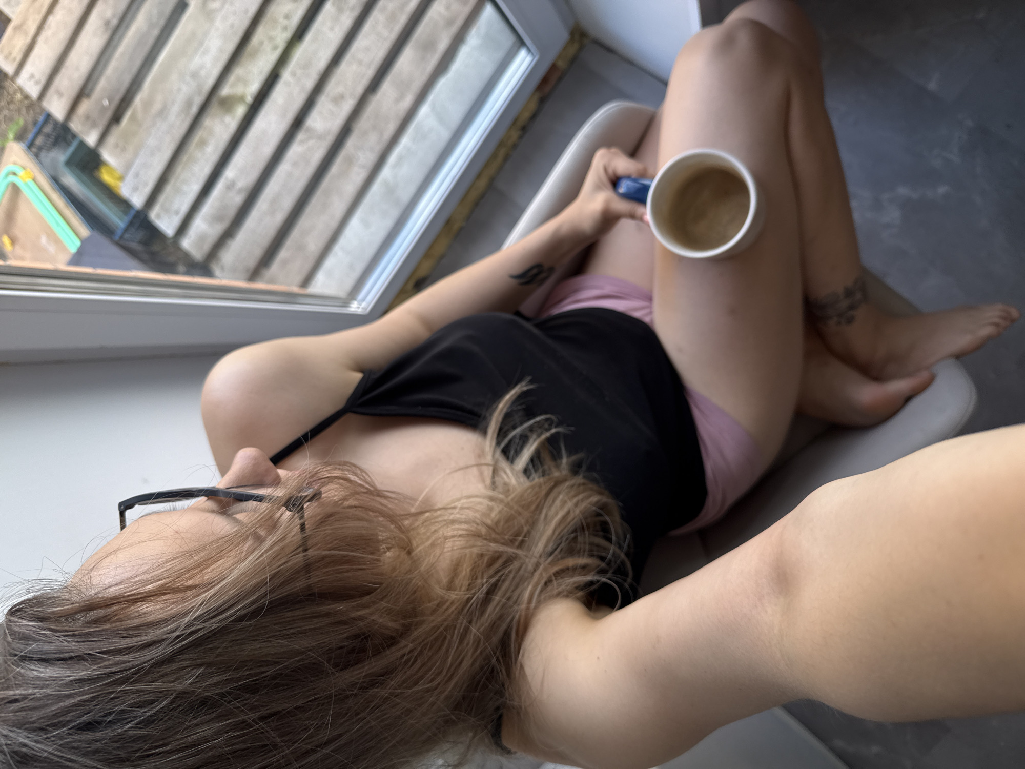 An overhead, high-angle shot captures a person relaxing indoors next to a large window. The individual, likely female, has long, light brown hair cascading around her head, and is wearing black-rimmed glasses. She is dressed in a black tank top and light pink underwear or shorts, and is seated on a light-colored chair with her legs bent and crossed.
In her right hand, she holds a white ceramic mug with a blue handle, containing a light brown beverage, possibly coffee or tea, which rests on her bent right knee. Tattoos are visible on her left forearm, appearing as stylized wings, and an indistinct pattern or text is tattooed on her right ankle/lower leg.
Through the window on the left, an outdoor scene is visible, featuring wooden decking or slatted surfaces. Hints of green and yellow objects are partially seen beyond the decking, suggesting a garden or outdoor area. The interior floor appears to be a dark, possibly tiled or concrete surface. The lighting indicates it is daytime, providing a serene ambiance for the person's relaxed posture. This tranquil scene is set in Chaikovsky, Russia.