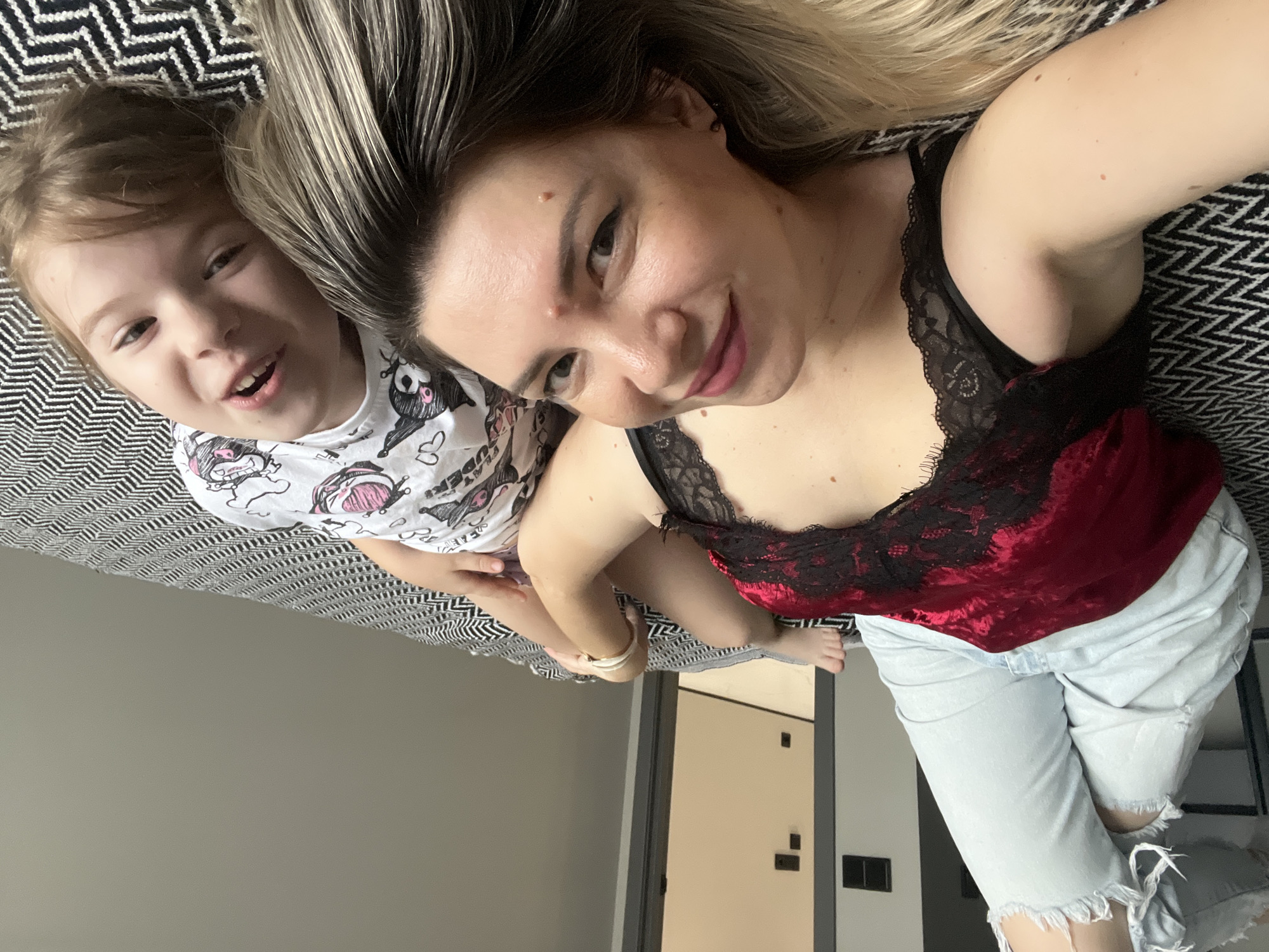 A young child with blond hair and a woman with long blond hair are lying down on a black and white patterned surface. The child is wearing a white t-shirt with cartoon characters printed on it. The woman is wearing a red and black lace camisole and ripped light blue jeans. They are both smiling and looking at the camera. The scene appears to be indoors, possibly in a bedroom, as suggested by the bed and a doorway in the background. The overall mood is cheerful and intimate, indicating a candid moment between the two subjects. The image appears to have been taken during the day.
